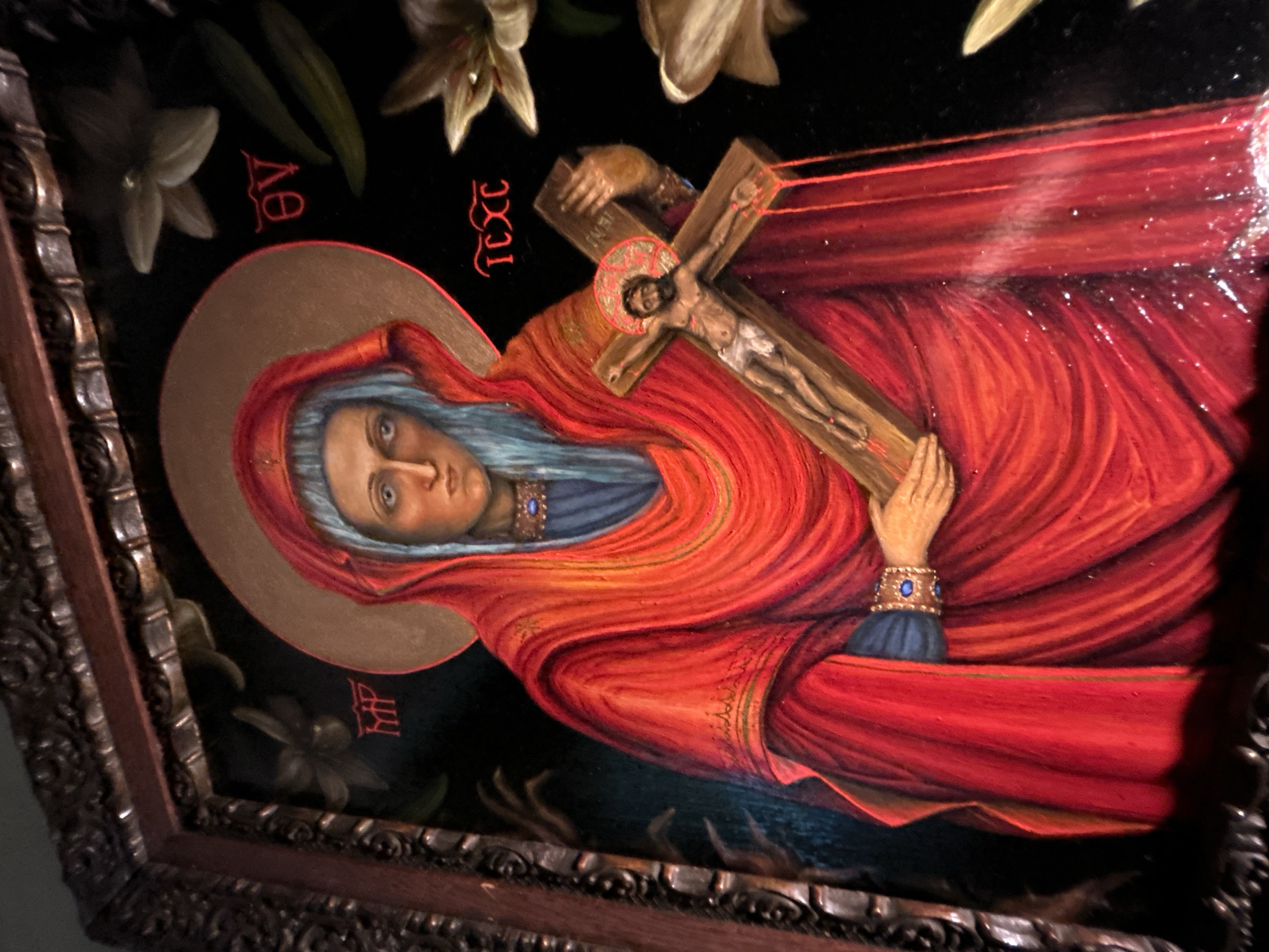 The image shows a framed painting of the Virgin Mary holding the crucified Christ. The painting is a religious icon style with gold halo and red robes. The background is dark, with some lilies. The frame is ornate and dark wood. There is no information to suggest this image was captured in Cape Coral, United States of America. Therefore, I cannot include a location.