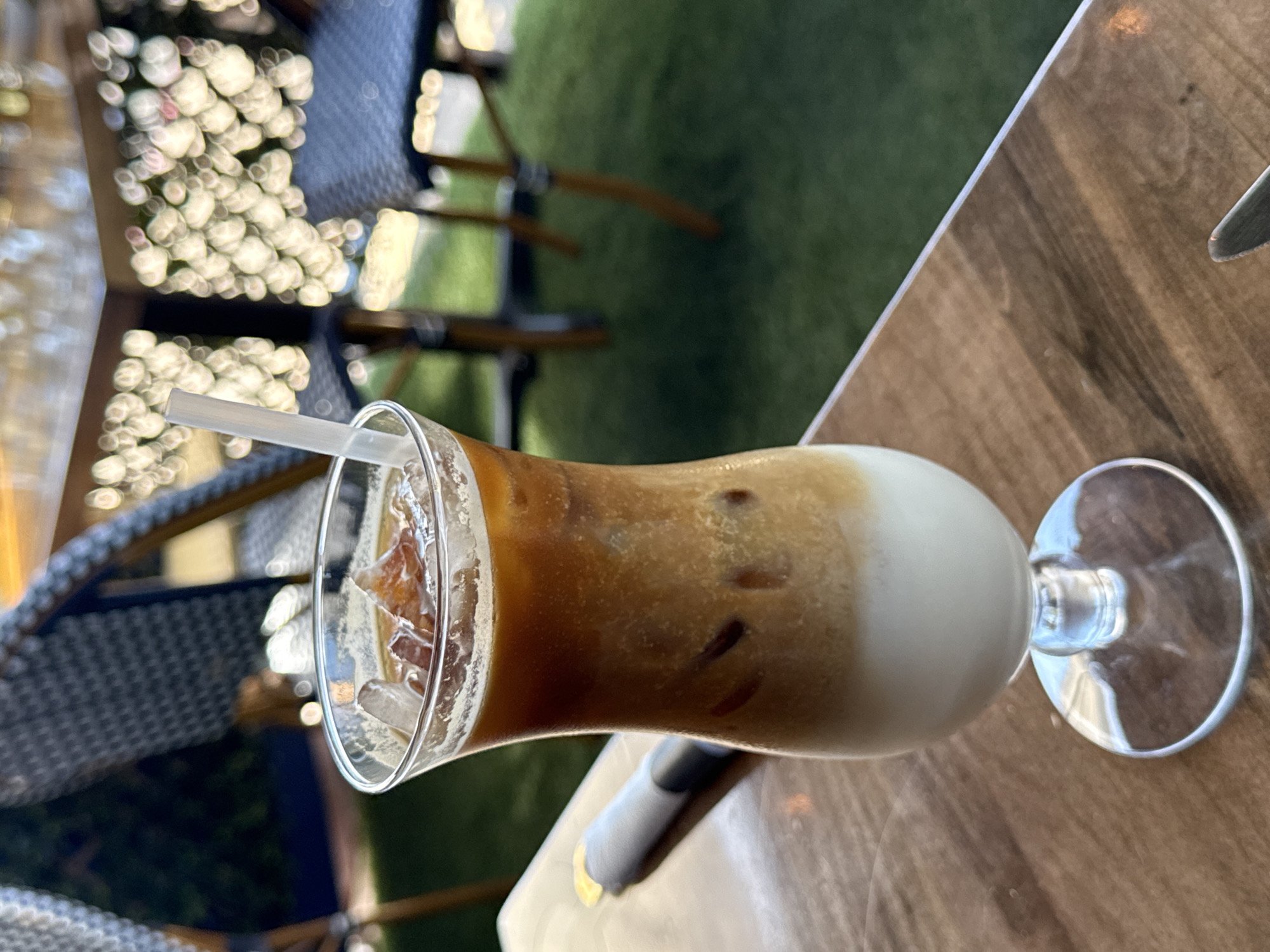 The image shows a close-up of a glass of iced coffee on a wooden table, with a straw in the glass. The background includes outdoor seating with chairs and a blurred green area. The location is Chula Vista, United States of America.