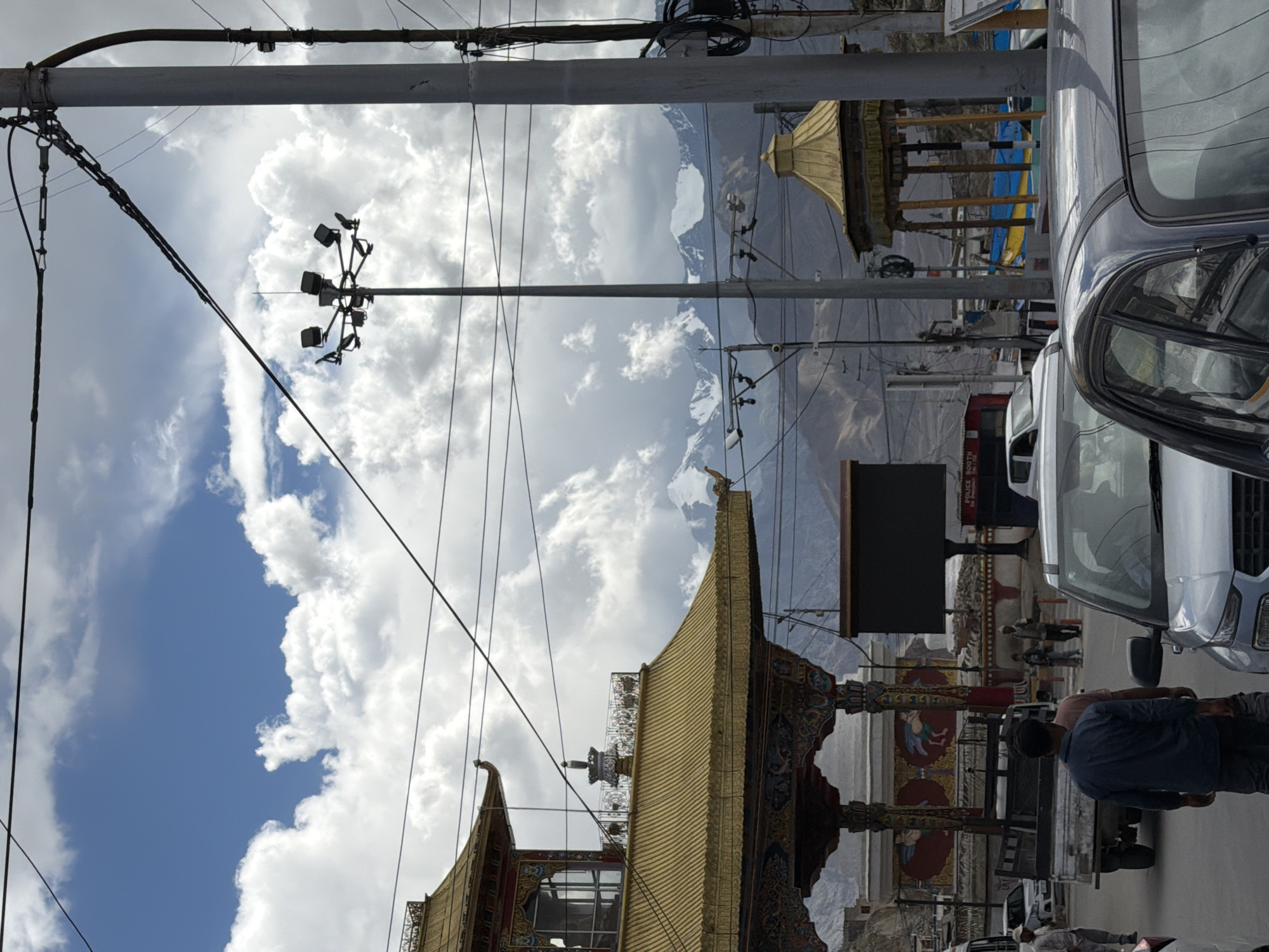 The image depicts a street scene in Leh, India, characterized by a mix of traditional architecture and modern infrastructure. Dominating the left side is a multi-tiered building with a golden, pagoda-style roof, adorned with intricate carvings and paintings. This structure appears to be a significant landmark, possibly a temple or a heritage building. In the background, imposing snow-capped mountains rise under a sky filled with dramatic, cumulus clouds.
The foreground shows a paved road with several vehicles, including cars and a small truck. People are visible walking on the roadside and near the vehicles, suggesting a bustling urban environment. Overhead, a complex network of power lines and utility poles crisscrosses the sky, indicating the presence of electricity and communication infrastructure. A tall lamppost with multiple lights stands prominently in the mid-ground, and a large digital billboard is also visible. The overall impression is of a vibrant street in a mountainous region, where cultural heritage coexists with the daily life of the city.
