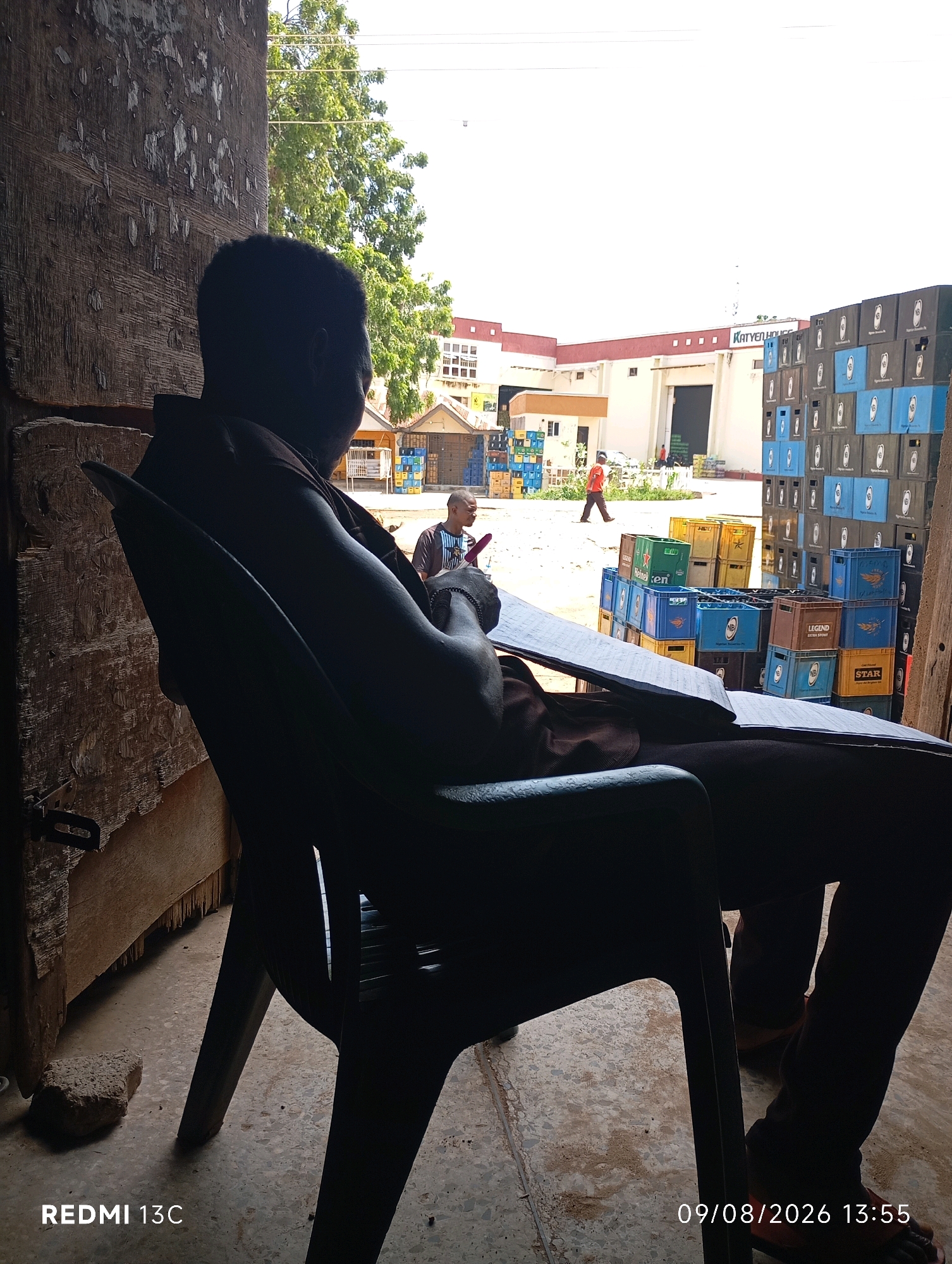 A person sits in a dark plastic chair, silhouetted against an open doorway looking out onto a street. The person is facing away from the camera, looking towards a building with a sign that reads "KATYEM HOUSE." To the right of the building are stacks of blue and black crates. In the foreground, on the ground, is a piece of concrete. The doorway appears to be made of rough, weathered wood. The floor is concrete. The time stamp on the image reads "09/08/2026 13:55."