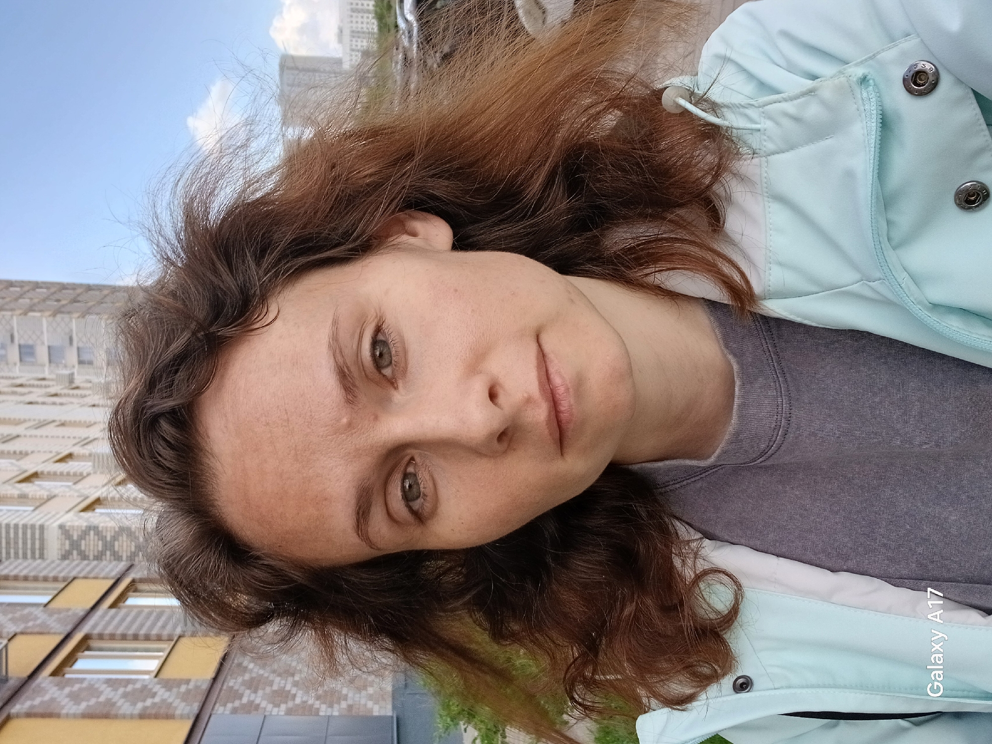 The image is a bright, daylight selfie of a young adult woman taken outdoors in an urban environment, likely in Yekaterinburg, Russia. The woman has long, wavy brown hair with reddish highlights, fair skin with visible freckles, and green/hazel eyes. She wears a light blue or mint green jacket with silver snap buttons and a zipper detail, over a grey long-sleeved shirt or sweatshirt. Her expression is neutral and calm.
The photograph is taken at a significant tilt, with the woman's head leaning towards the left side of the frame. In the background, there's a modern building featuring a patterned facade of light-colored bricks and yellowish accent panels, with visible rectangular windows. Taller buildings or skyscrapers can be seen further in the distance, silhouetted against a clear blue sky with some white clouds. A hint of greenery is visible in the lower right background. The lighting suggests it is either late morning or early afternoon. A "Galaxy A17" watermark is present in the bottom right corner of the image.