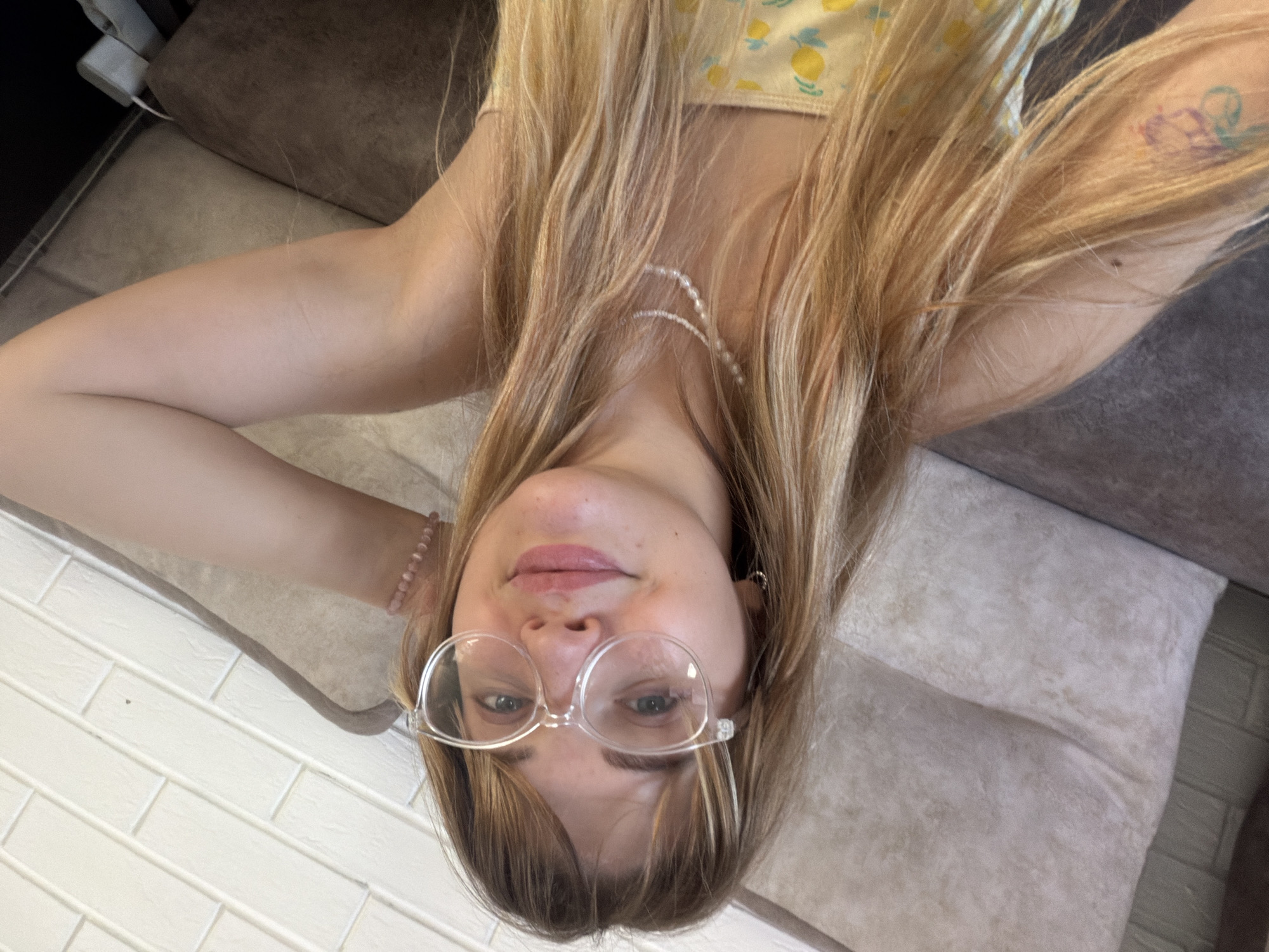 A young person with long blonde hair is reclining on a light-colored sofa, wearing glasses and a yellow top with fruit patterns. The location appears to be an indoor setting, potentially a home or apartment, with a section of white tiled flooring visible in the lower left. The person has a light skin tone and is wearing a pearl necklace and a pink bracelet. The person is positioned in front of a sofa in Большая Шильна, Russia.