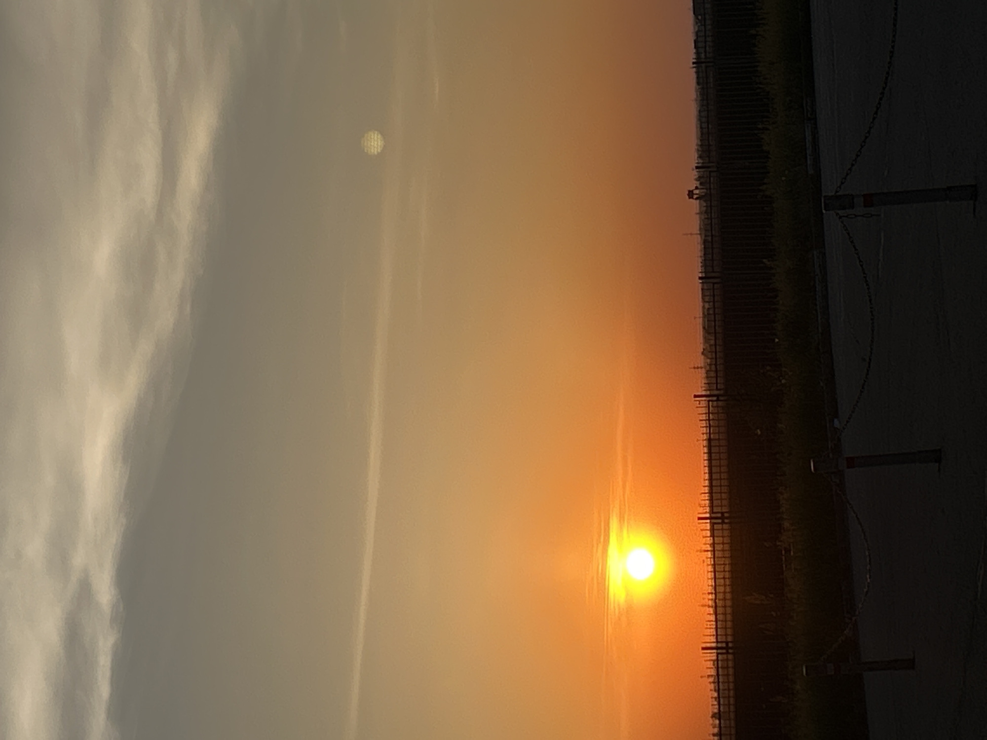The image shows a sunset or sunrise with the sun low on the horizon, appearing as a bright, orange-yellow orb. To the left of the sun, the sky transitions into a lighter, hazy, orange-yellow color. Above the horizon line, the sky is lighter and more muted. On the left side of the image, there are visible white clouds. On the right side of the image, there is a dark fence and some vegetation, partially obscured by shadow. In the lower-right corner, there is a portion of a paved surface and part of what appears to be a chain-link barrier. Another dimmer solar disc is visible above and left of the primary sun image.
The location is a city of Unknown, Unknown.