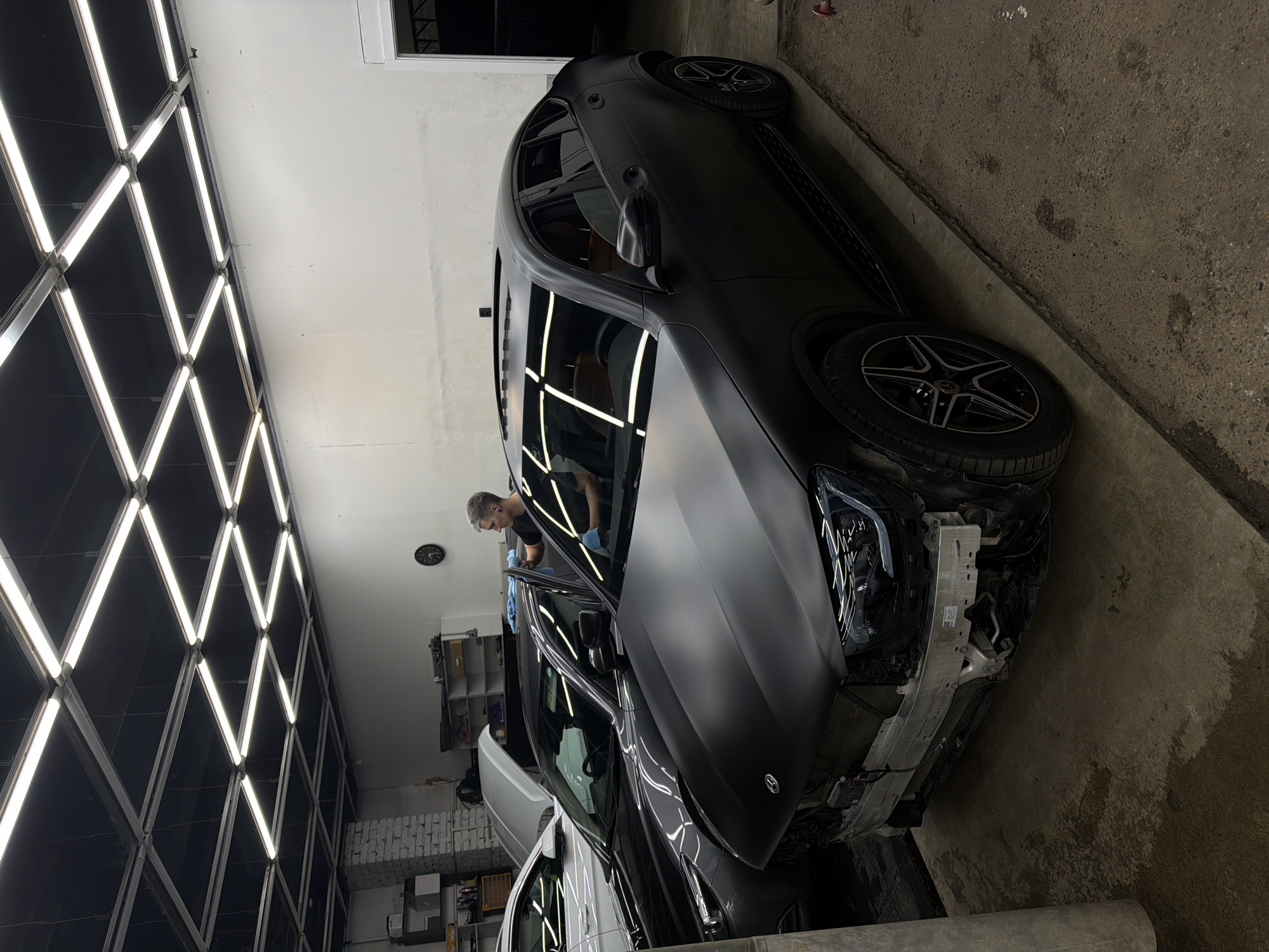 In an indoor automotive workshop, a matte black Mercedes-Benz coupe SUV, partially disassembled at the front bumper, is the primary focus. The vehicle has a panoramic sunroof and light-colored leather interior visible through the windows. To the left of this vehicle, a dark grey sedan with its hood open is parked. Further to the left, a white car is partially visible. A man, wearing blue gloves and a black t-shirt, is standing between the sedan and the SUV, appearing to be cleaning or applying something to the driver's side window of the sedan. The workshop has a concrete floor and white walls. The ceiling is fitted with a grid of fluorescent light fixtures. On the wall, a round clock is visible. Shelving units containing various automotive supplies are present in the background. The overall environment suggests ongoing automotive detailing or repair work. The location is identified as Krasnoyarsk, Russia.