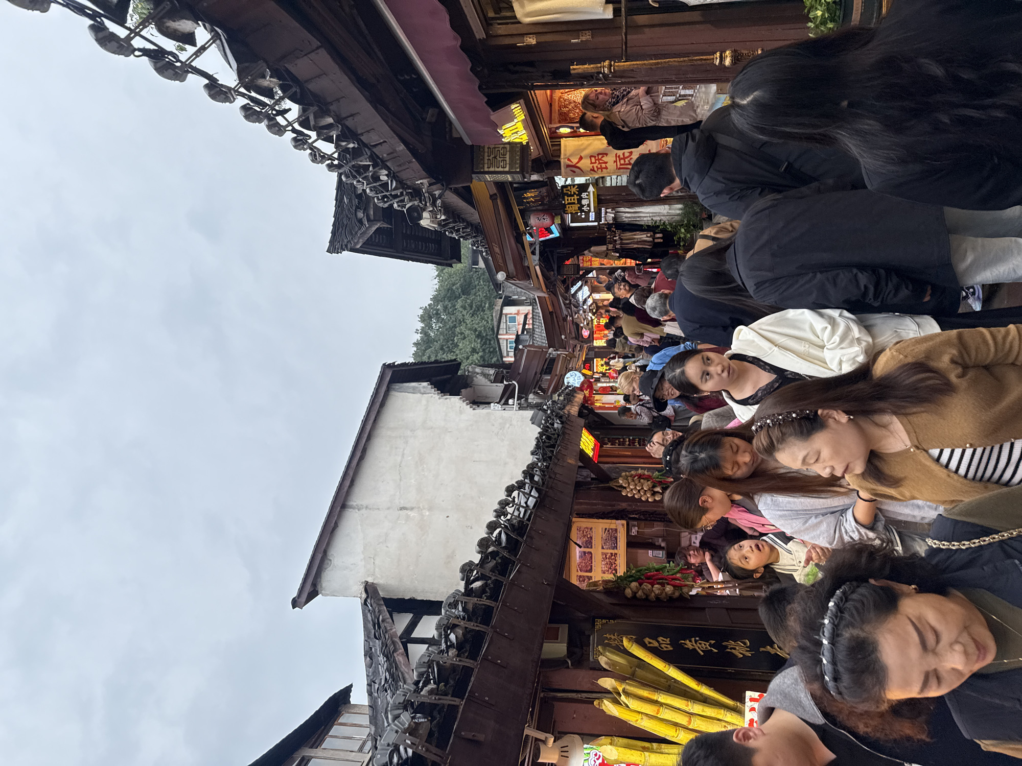 The image depicts a bustling street scene in the Shapingba District of Chongqing, China. A dense crowd of people of various ages and attire fills a narrow pedestrian walkway lined with traditional Chinese architecture. Buildings with dark wooden beams and tiled roofs are visible, adorned with ornate details and hanging decorations. Brightly lit storefronts display various goods, including fruits, vegetables, and food items. The overall atmosphere is lively and crowded, with a mix of locals and tourists likely present. The sky appears overcast, casting a soft light on the scene.