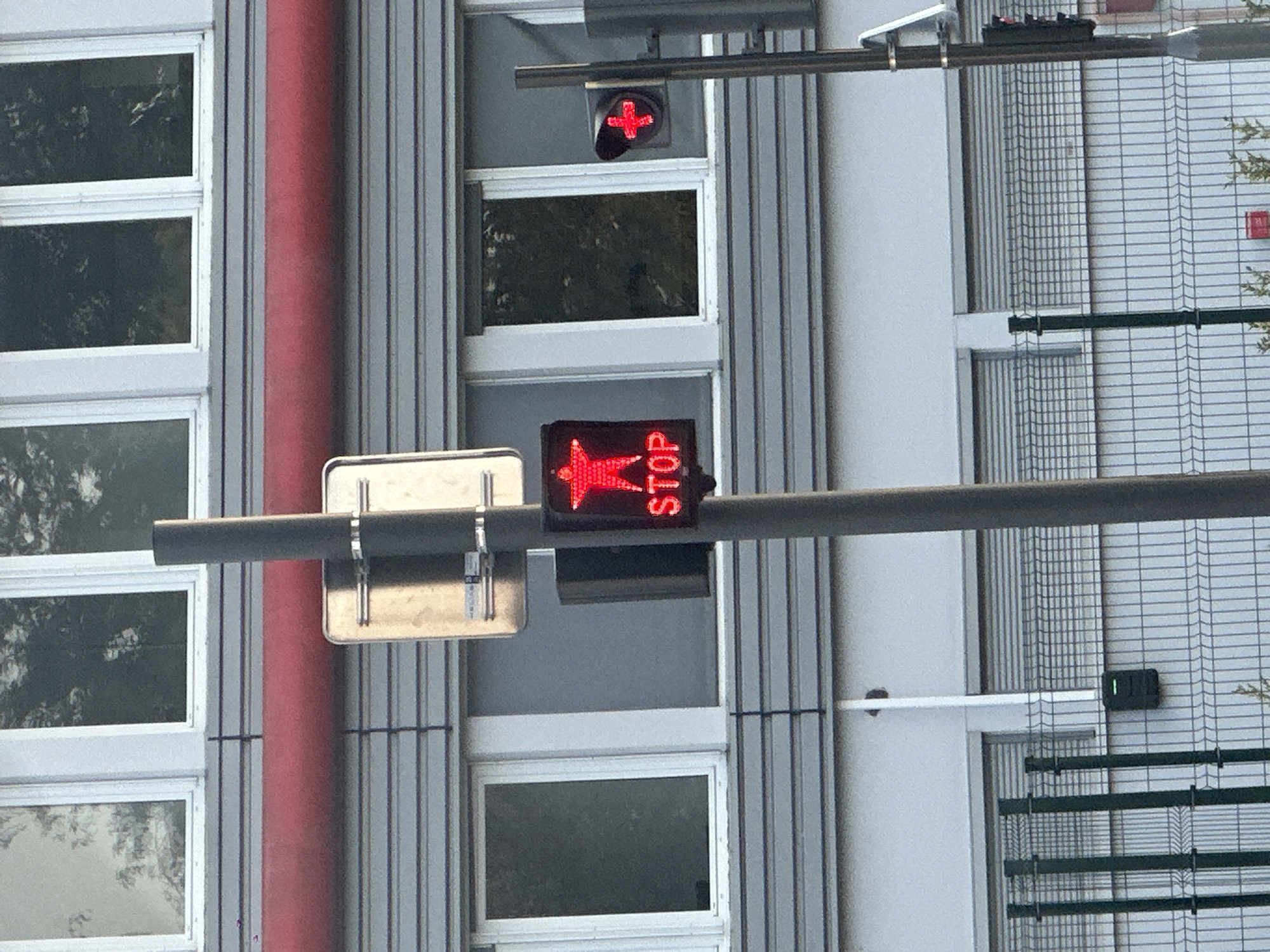 The image depicts a street scene in Eckbolsheim, France. In the foreground, a traffic light pole with a red illuminated pedestrian signal showing a star and the word "STOP" is visible. To the right of this signal, another traffic light is visible, also illuminated red with a cross symbol. Behind the traffic lights, a grey building with horizontal grey siding and several windows is present. A thick red pipe runs horizontally across the building facade above the windows. A metal fence runs along the bottom of the image, with a dark green electronic box attached to a white conduit on the building wall behind it. The overall scene suggests an urban environment, likely a street crossing or intersection.