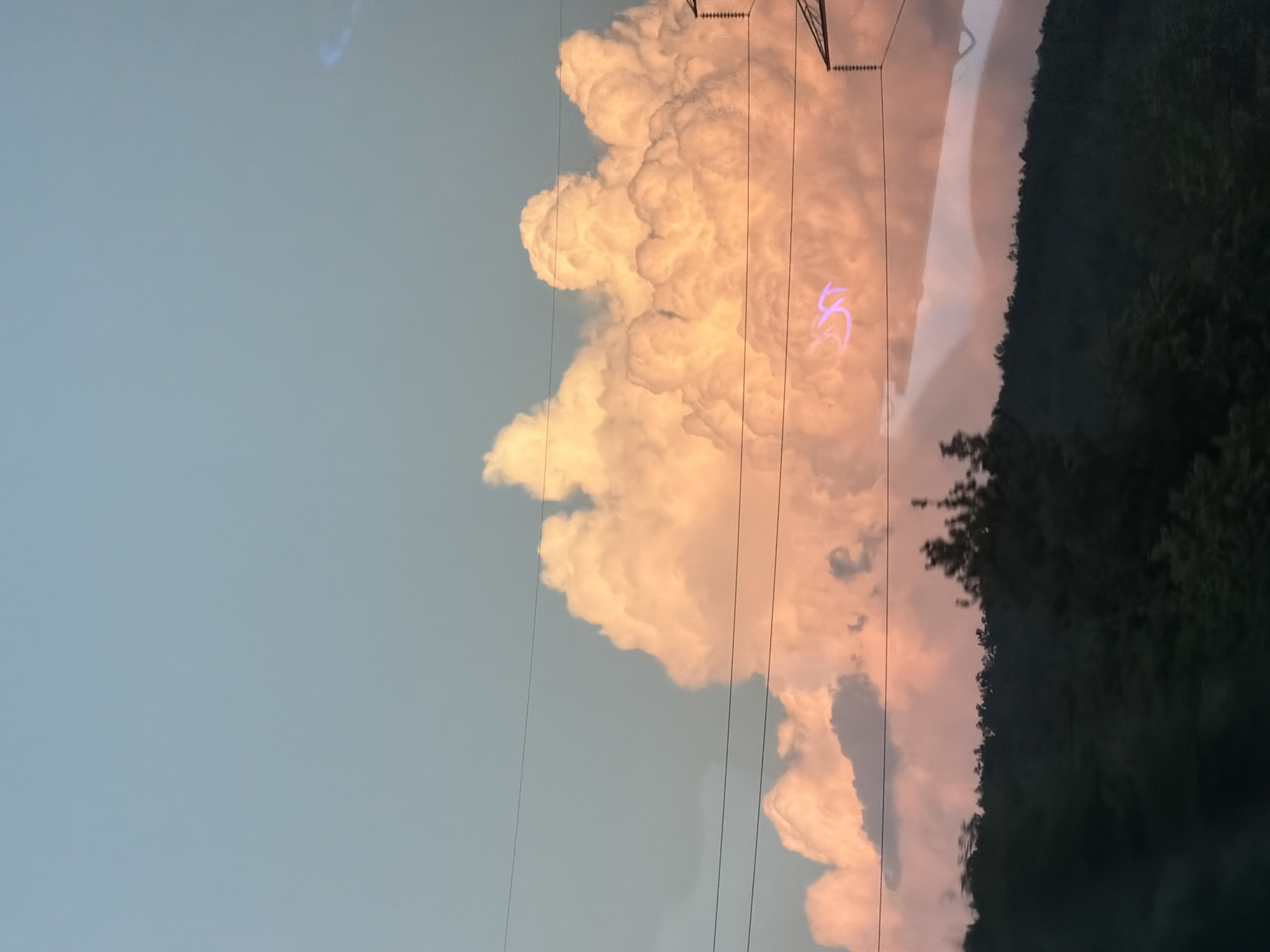 A sky of pale blue and soft pink is dominated by voluminous, illuminated clouds that appear to be catching the light of a setting sun. In the foreground, a dark, hilly landscape is covered with trees, their silhouettes creating a natural horizon line. Several power lines crisscross the frame, adding a man-made element to the scene. A faint, purple, abstract shape is visible in the sky, possibly an artifact of the camera or a light anomaly. The image is a landscape captured from a moving vehicle, as evidenced by the slight blur of the foreground trees. The specific location in Rajana, Bulgaria, is not discernible from the visual content of the image alone.