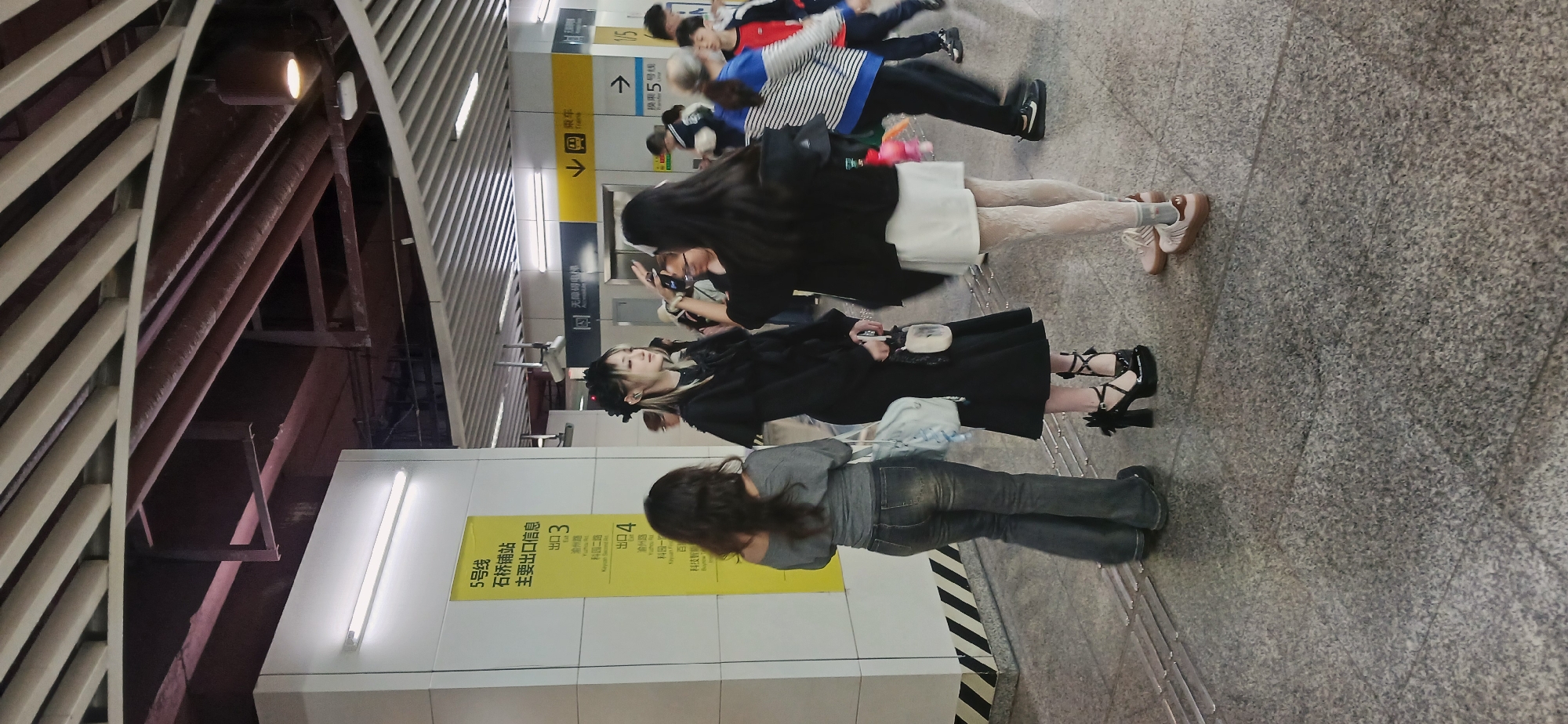 The image depicts a subway station in Jiulongpo District, China, as indicated by the sign for the "Shuqiaopu" station. Several people are present, with some individuals facing the camera and others facing away. A group of people can be seen closer to the camera. One individual is wearing a black coat, a white skirt, patterned leggings, and light-colored shoes. Another person is wearing a black dress and black heels with decorative bows. Another individual is wearing a grey top and jeans. One person is using a cell phone. In the background, there are signs with arrows indicating directions for train lines and stations. The station has a modern architectural design with overhead structures and lighting. The floor is grey and appears to be made of stone tiles. The lighting seems to be a combination of natural and artificial. The scene appears to be a candid shot of people waiting in the subway station.