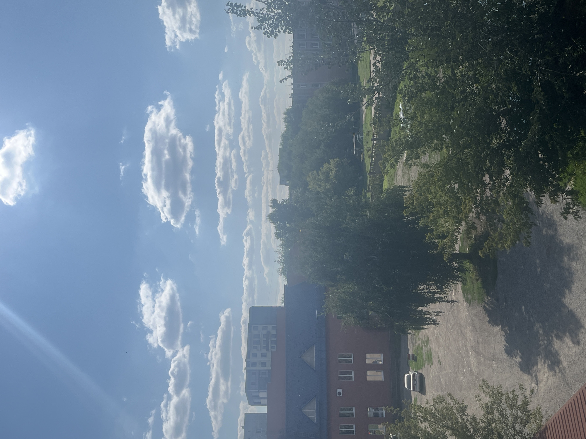 A daytime, medium-shot photograph captures a view of buildings and trees under a partially cloudy blue sky in Oskemen, Kazakhstan. In the foreground, the upper portion of a red-tiled roof is visible. To the left and right, the dense foliage of trees partially obscures the view. A gravel parking lot is situated in the midground, with a white car parked at an angle on the left side. Behind the parking lot, a two-story brick building with a dark roof is prominent, featuring multiple windows and two triangular architectural elements on the roof. Further in the background, several other buildings, including multi-story residential structures, are visible, interspersed with trees. The sky is a bright blue, with scattered white, puffy clouds and a visible sun glare in the upper left corner.