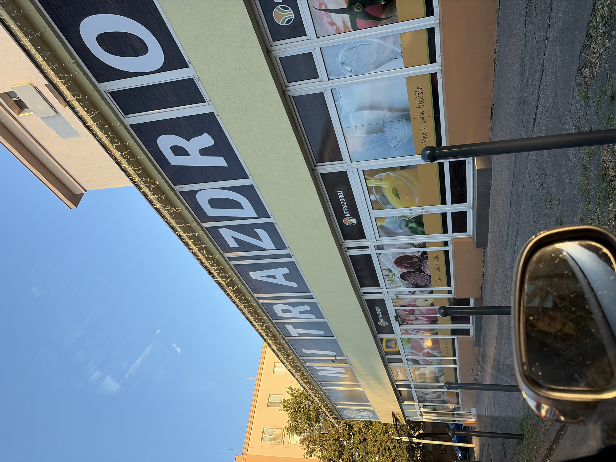 The image captures a storefront in Nitra, Slovakia, bearing the prominent signage "NITRAZDROJ". The building appears to be a commercial establishment, likely a supermarket or grocery store, indicated by the images displayed on its windows. These images depict various food items, including what appears to be cured meats and dairy products, as well as a pitcher of juice. The storefront features large glass windows and a yellow facade. Above the windows, large white letters spell out "NITRAZDROJ" against a dark background. Decorative string lights are visible along the top edge of the storefront. In the foreground, a car's side mirror is visible, reflecting a person taking a photograph. The street in front of the store is paved, with bollards and a bicycle rack present. The sky is clear and blue, suggesting a daytime setting. The overall scene depicts an urban commercial environment in Nitra.