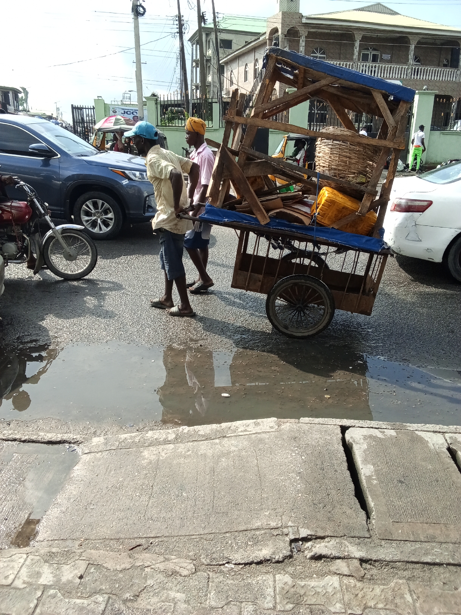 The image depicts a vibrant street scene in Ado, Nigeria, under bright daylight. Two men are seen actively pushing a heavily loaded, two-wheeled wooden cart along the asphalt road. The cart is piled with items including a large woven basket, a yellow plastic jerrycan, and various wooden pieces, partially covered by a blue tarp. The man in front wears a light yellow shirt, blue cap, and shorts, while his companion behind him has a pink shirt and a yellow headwrap.
The road features a large puddle of standing water that reflects the men and the cart. Surrounding them is moderate traffic, including a blue SUV, the front wheel of a red motorcycle, and the rear of a white sedan with visible damage. In the background, a yellow tricycle (keke Napep) can be seen. Buildings line the street, one featuring a green gate and another with multiple balconies. Overhead utility poles and wires are prominent against the clear sky.
The strong sunlight creates distinct shadows, indicating a sunny day, although the puddle suggests previous rain or poor drainage. On a building or sign in the background, the text "JCC" and partially "READY TO SERVE" are visible. The scene captures an everyday moment of manual transport and urban activity.
