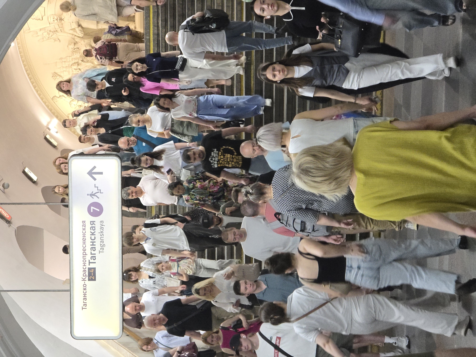 The image depicts a crowded staircase in the Taganskaya metro station in Moscow, Russia. Numerous people of various ages and appearances are walking up and down the stairs. A sign indicating directions to the Tagansko-Krasnopresnenskaya line (lines 2-4) and Taganskaya station (line 7) is visible. There is a mural on the wall near the top of the staircase. The setting appears to be a busy public transportation hub.