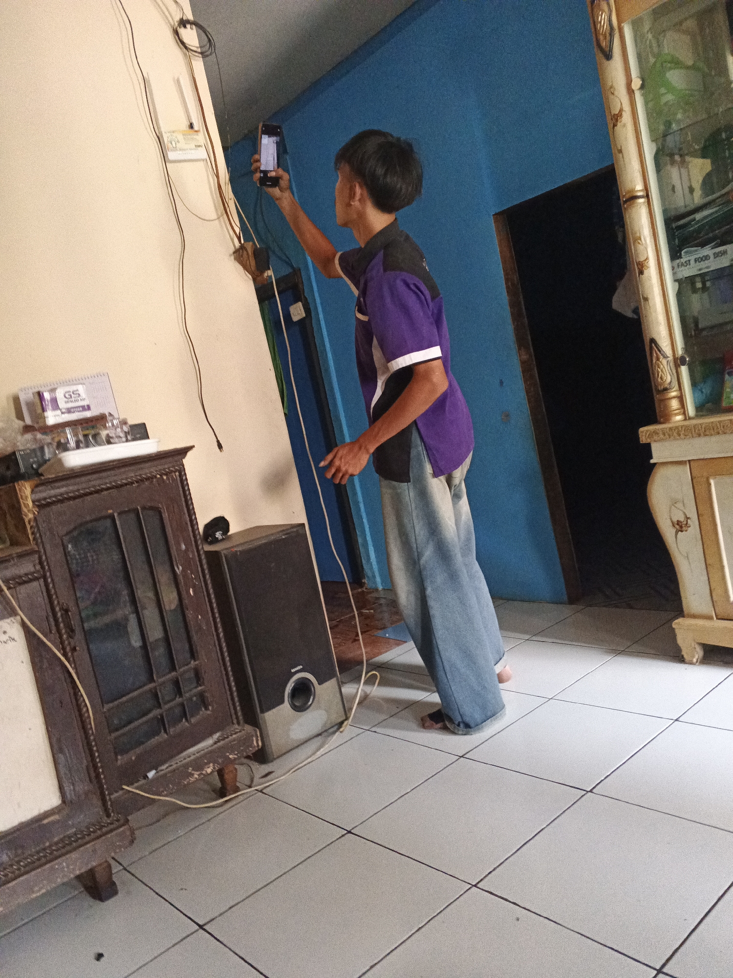 A person is standing in a room and taking a selfie with their phone. The room has blue walls, white tiled floors, and a wooden cabinet. There is a speaker next to the cabinet. A router is mounted on the wall above the cabinet. The person is wearing a purple and black shirt and blue jeans. The person's feet are bare.