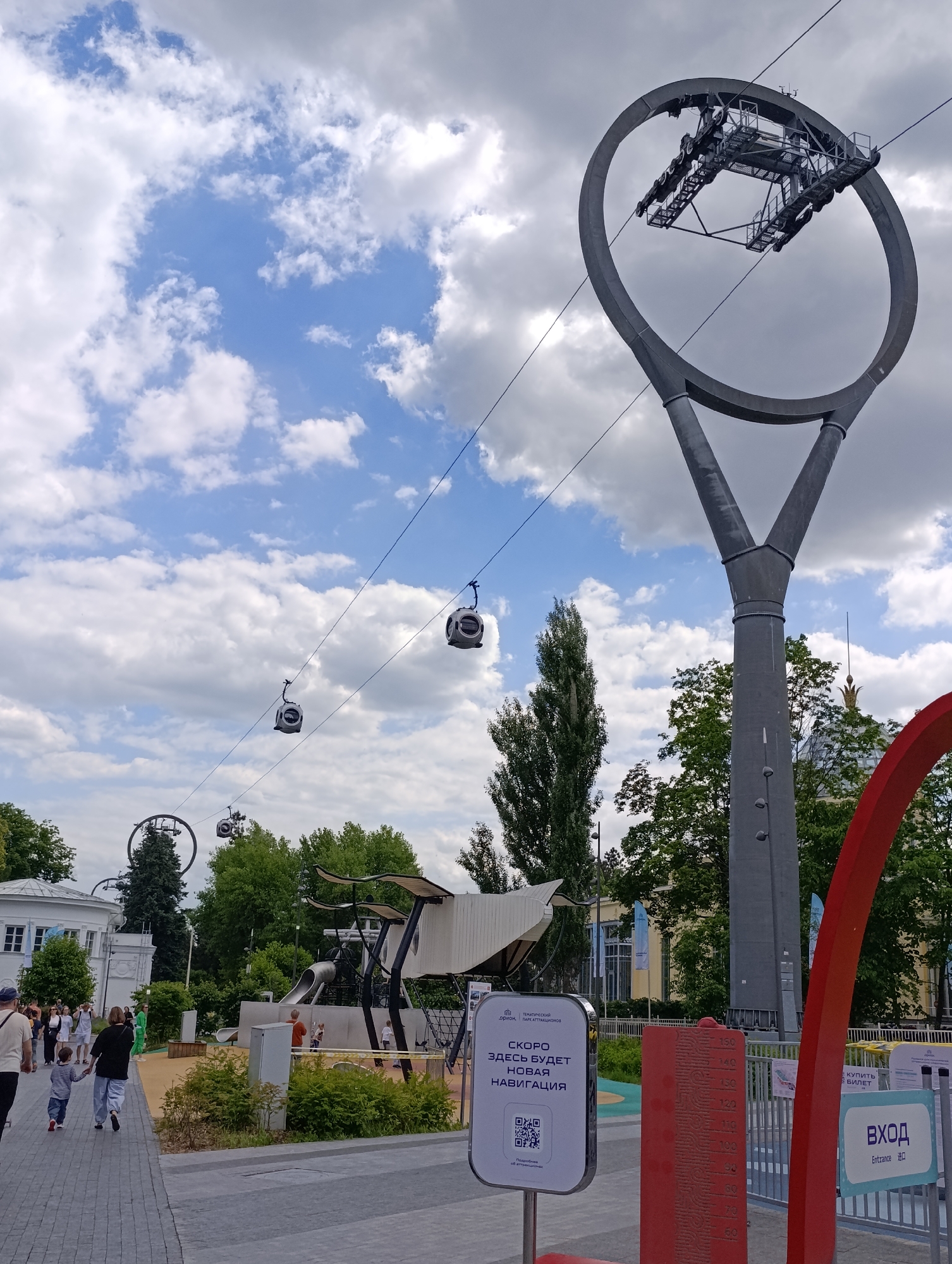 This daytime image captures a vibrant recreational area in Moscow, Russia, featuring a modern cable car system operating under a partly cloudy sky. The scene depicts a paved pathway populated by several individuals, including adults and children, strolling casually.
The dominant features are the sleek, gray cable car system, characterized by multiple individual gondolas suspended along cables, and two large, circular support pylons. One pylon, prominently in the right foreground, has a distinctive Y-shaped base. In the middle distance, another similar pylon is visible. Below the moving gondolas, a modern playground structure with slides and climbing elements is situated amidst green trees and bushes. A white building with traditional architecture is seen on the left, while another building with a golden spire is partially visible through the foliage on the right.
On the ground level, near the pathway, a red archway marks what appears to be an entrance. Adjacent to it is a red height-measuring stick with numbers from 50 to 150 cm. A white information sign in the foreground displays Russian text: "ТЕМАТИЧЕСКИЙ ПАРК АТТРАКЦИОНОВ" (THEMATIC AMUSEMENT PARK) and "СКОРО ЗДЕСЬ БУДЕТ НОВАЯ НАВИГАЦИЯ" (SOON THERE WILL BE NEW NAVIGATION HERE), accompanied by a QR code and "Подробнее об аттракционах" (More about the attractions). A partially visible sign to the right states "Купить БИЛЕТ" (Buy TICKET) and "Вход Entrance 进口" (Entrance in Russian, English, and Chinese), indicating a public, likely international, attraction. The overall atmosphere suggests a pleasant day out at a park or entertainment complex.