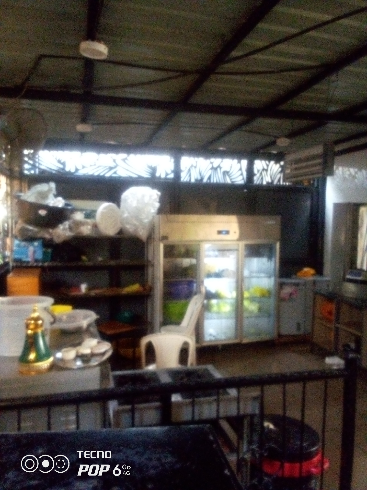 The image shows an indoor scene, likely a food service establishment, possibly in Howayi, Nigeria. The environment is dimly lit, with a corrugated metal roof supported by a dark metal frame. Two ceiling fans are visible. In the foreground, a black metal railing separates the viewer from the interior. Behind the railing, a dark trash bin with red accents is visible.
The interior features shelves stocked with various items, including what appear to be containers and possibly food items. A large, illuminated commercial refrigerator or display cooler is prominently positioned, filled with what looks like produce or prepared food items. A white plastic chair is placed in front of the cooler. On a counter to the left, there is a decorative golden and green dispenser or kettle, along with a tray holding small cups, suggesting a beverage service. Another large white container is also on the counter. In the background, there are further shelves and what could be a counter or preparation area. A decorative screen with a floral pattern is visible in the upper background, possibly as part of the wall or window treatment. The overall impression is of a functional, possibly casual, food service area.