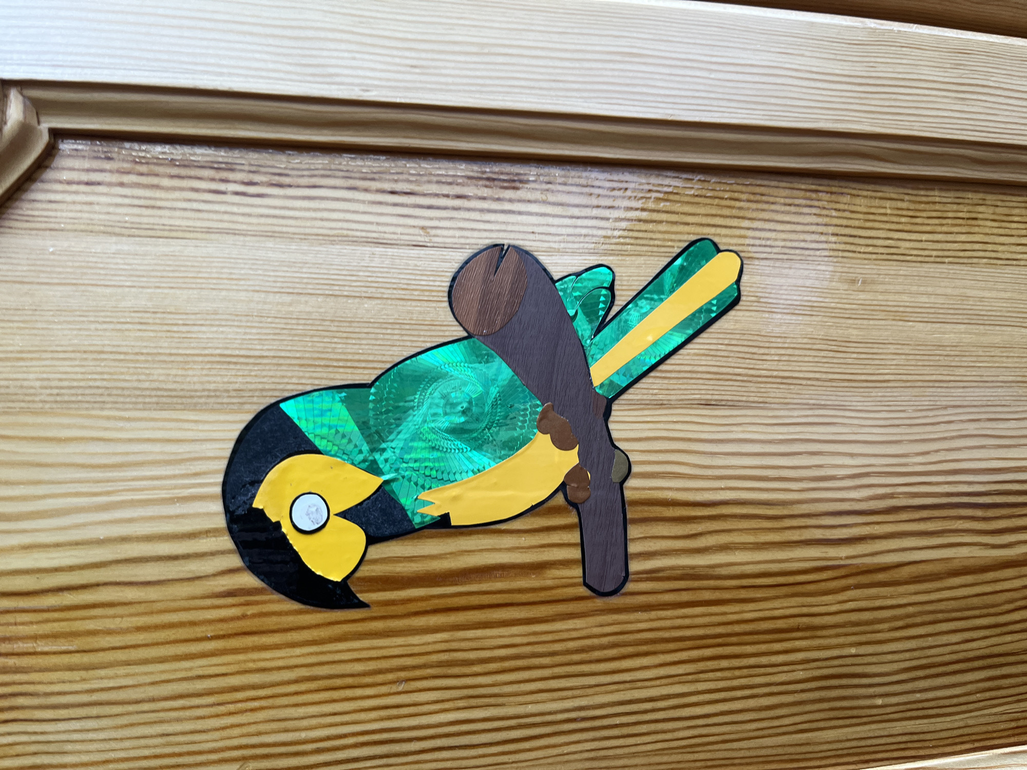 A colorful, cutout parrot decoration is affixed to a wooden surface, possibly a door or cabinet. The parrot is depicted in profile, perched on a brown branch. It has a black head with a yellow face and a white eye. Its body is primarily green with a holographic, patterned sheen, and it has a yellow beak and tail feathers accented with green stripes. The wooden surface features prominent vertical grain lines and a light, honey-brown finish. To the right of the main wooden surface, a vertical seam indicates a panel or edge, with a lighter-toned wood visible on the far right. The lighting suggests an indoor environment, possibly a home. The location is Unknown, Unknown.