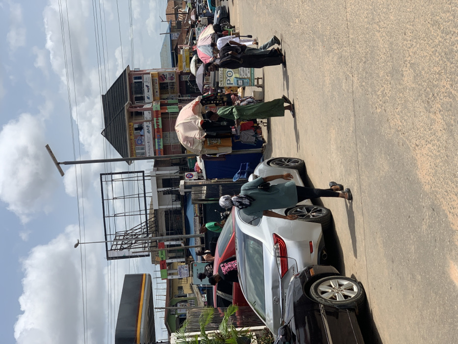 The image shows a street scene in Akure, Nigeria, at the coordinates 7.252 latitude and 5.198 longitude. There are several people walking on the street, some of whom appear to be shopping at various roadside stalls. A silver car is parked in the middle of the street, and a black car is parked on the left side of the street. The street is paved and dusty. In the background, there are several buildings, including a two-story building with a sign that reads "New Boutique." There is also a large billboard that is blank. The sky is partly cloudy. The people in the foreground are wearing various clothing. Some of them are carrying bags or purses. One person is wearing a headscarf. The environment is busy and chaotic, with traffic, pedestrians, and vendors all vying for space.