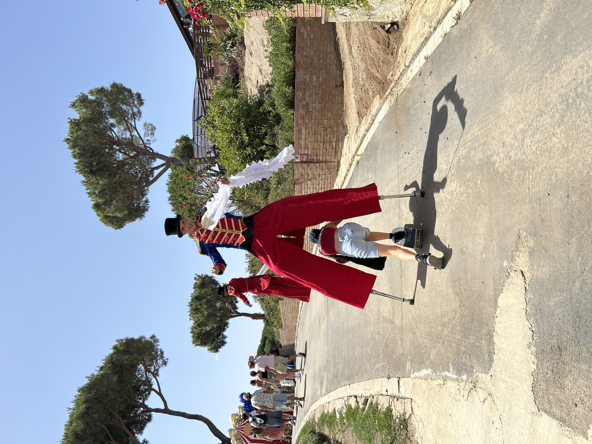 In Sant Vicenç De Montalt, Spain, on a sunlit day, a stilt walker in a red and blue circus-style costume, complete with a black top hat, stands on tall stilts. The performer holds a white fabric in their right hand. In front of the stilt walker, a person on a skateboard rides in the same direction, their back to the camera, wearing a red top and denim shorts. The skateboarder is positioned between the legs of the stilt walker.
The scene unfolds on a paved pathway with a slight incline. To the left of the pathway, there is a grassy verge and beyond that, a line of trees, some of which appear to be pine trees. Further in the background, other people are walking, some carrying umbrellas, suggesting a public gathering or event. To the right of the pathway, a brick wall runs along the side of a gently sloping, vegetated hillside with more trees and some flowering plants. A building with a wooden balcony is visible at the top of the hill. The sky is clear and blue. The overall atmosphere appears festive and outdoors.
