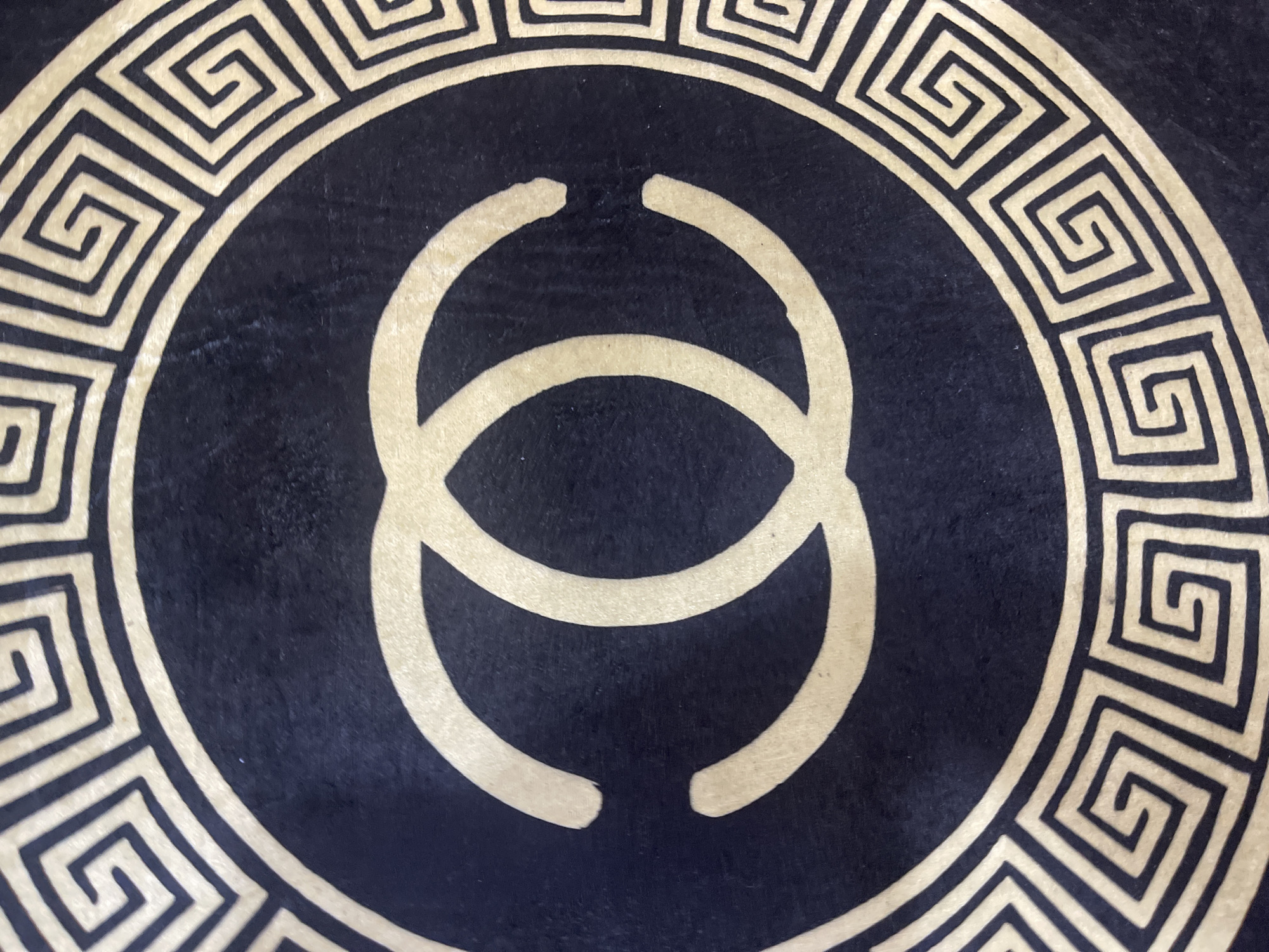 The image displays a close-up view of a dark circular pattern, likely on a fabric or rug. The pattern features a prominent golden interlocking "CC" logo in the center, reminiscent of the Chanel brand. Encircling the logo is a decorative border composed of a Greek key or meander pattern, also in golden lines against the dark background. The texture of the dark material appears to be slightly velvety or fuzzy. The image is an artistic or decorative close-up, with no environmental context provided. There are no people, actions, or specific city locations visible in the image.