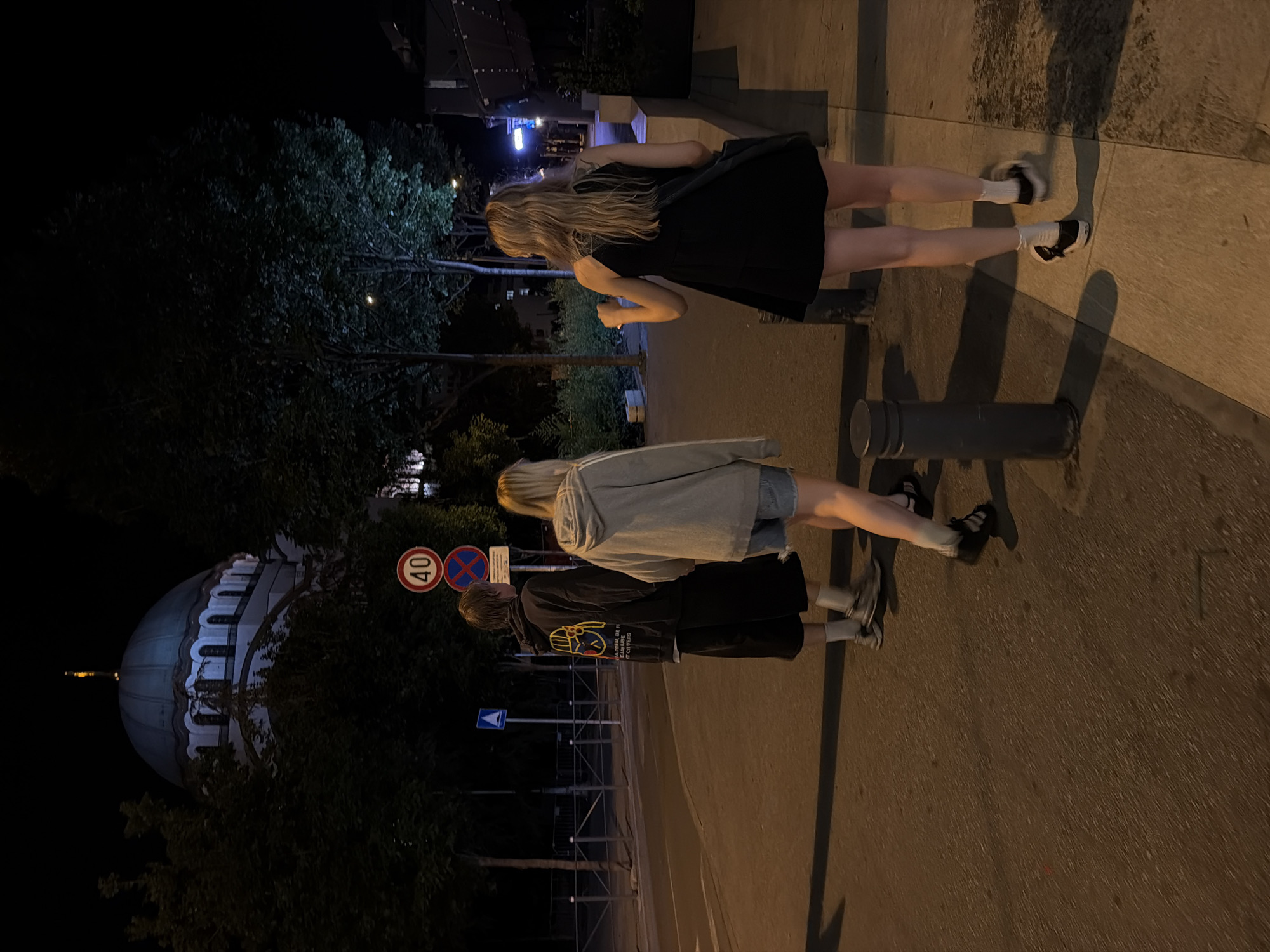 The image depicts a nighttime scene on a street in Vracar Urban Municipality, Serbia, with a prominent domed building in the background. Three individuals are walking away from the camera on a sidewalk adjacent to a street. The person closest to the camera on the right is wearing a black dress and white socks with black shoes. To their left, another person is wearing a grey hoodie and denim shorts. The furthest person, on the left, is wearing a dark hoodie with a graphic print on the back, black shorts, and white socks with grey sneakers. A traffic sign indicating a 40 km/h speed limit and another sign with a red circle and a diagonal bar are visible on the left side of the street, along with a blue directional sign. Trees line the street, and streetlights are casting shadows. The overall atmosphere is dark and urban.