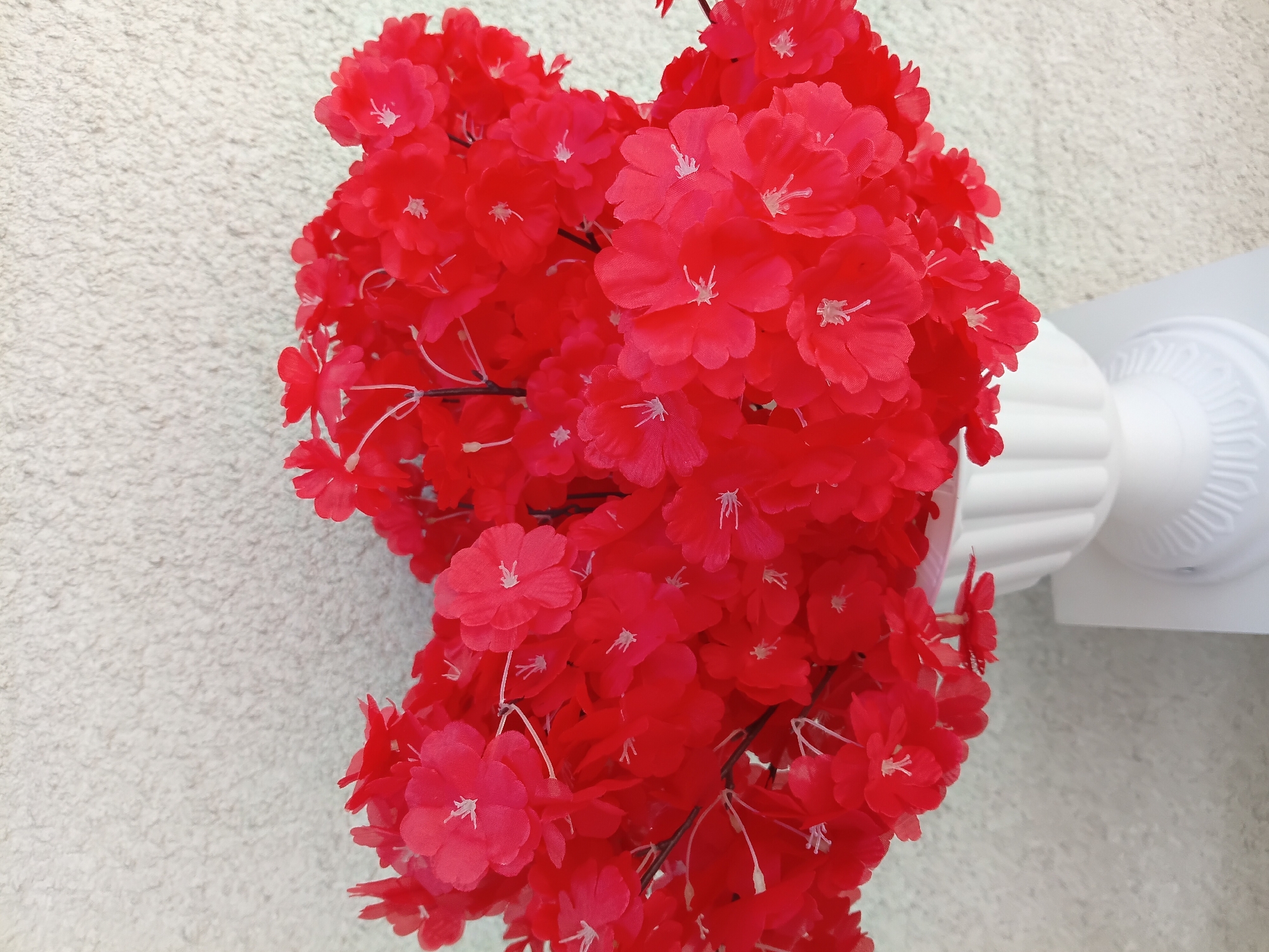 A bouquet of vibrant red artificial flowers is displayed in a white urn-shaped vase. The vase is placed on a white pedestal. The background is a textured, light-colored wall. The image is a close-up shot of the flowers and vase, with the wall filling the upper portion of the frame. There are no people or animals present in the image. The location in the city of Osh, Kyrgyzstan is not discernible from the visual information provided in the image.