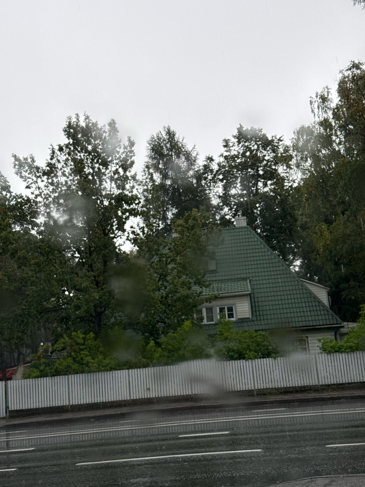The image depicts a residential house with a green tiled roof, surrounded by trees on a cloudy day. A white picket fence runs along the front of the property, separating it from a wet asphalt road with white lane markings, indicating it has recently rained. The scene is likely located in a suburban area of Tallinn, Estonia. There are no people or vehicles visible in the frame. The overall impression is one of a quiet, overcast day.
