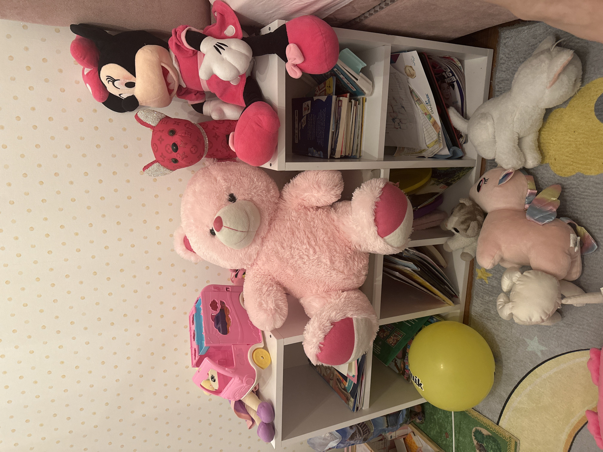 The image shows a white shelving unit filled with stuffed animals and books, resting on a patterned rug and in front of a polka-dot wall. The stuffed animals include Minnie Mouse, Mickey Mouse, a pink teddy bear, a red rabbit, a white cat, a pink pony with wings, and a smaller gray teddy bear. A yellow balloon is also visible near the bottom of the shelves. A pink plastic toy car is near the teddy bear. There is no evidence to indicate the location of the photo is in Esenyurt, Turkey.