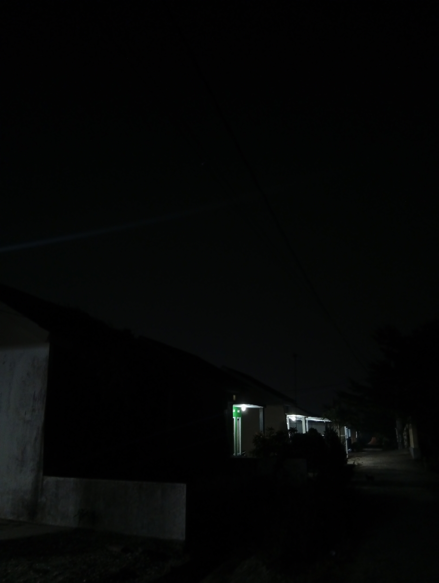 The image depicts a dark, nighttime scene of a residential street in Kualu, Indonesia. The foreground shows the edge of a building or structure, partially illuminated by ambient light. A sidewalk or pathway is visible in the mid-ground, lined with houses that have illuminated windows, some emitting a green glow. Trees and foliage obscure parts of the houses and the street. The sky is dark, with a thin, faint line that could be a light source or a wire stretching across it. The overall impression is one of quiet stillness, characteristic of a residential area at night.