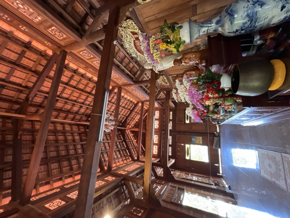 Intricate wooden beams create a geometric ceiling pattern above a room decorated with religious iconography. Statues, floral arrangements, and decorative plates adorn a surface, while windows provide natural light. The architecture and decorations suggest a temple or place of worship, showcasing detailed craftsmanship and spiritual significance. The structure emphasizes height and ornate design.