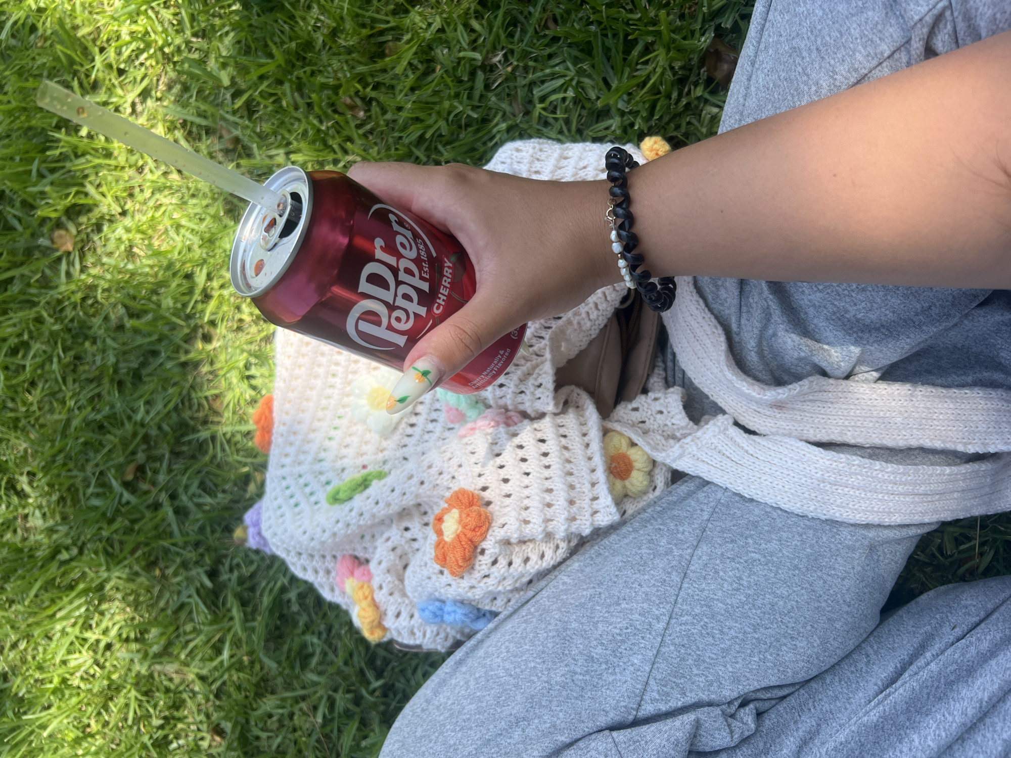 The image depicts a close-up, overhead view of a person's hand holding a can of Dr Pepper Cherry with a straw, resting on a crocheted garment with flower appliques. The person is wearing grey sweatpants and a bracelet with black and pearl beads. The scene is set on a grassy lawn, likely in Kiryat Bialik, Israel, suggesting an outdoor leisure activity. The lighting indicates a sunny day.