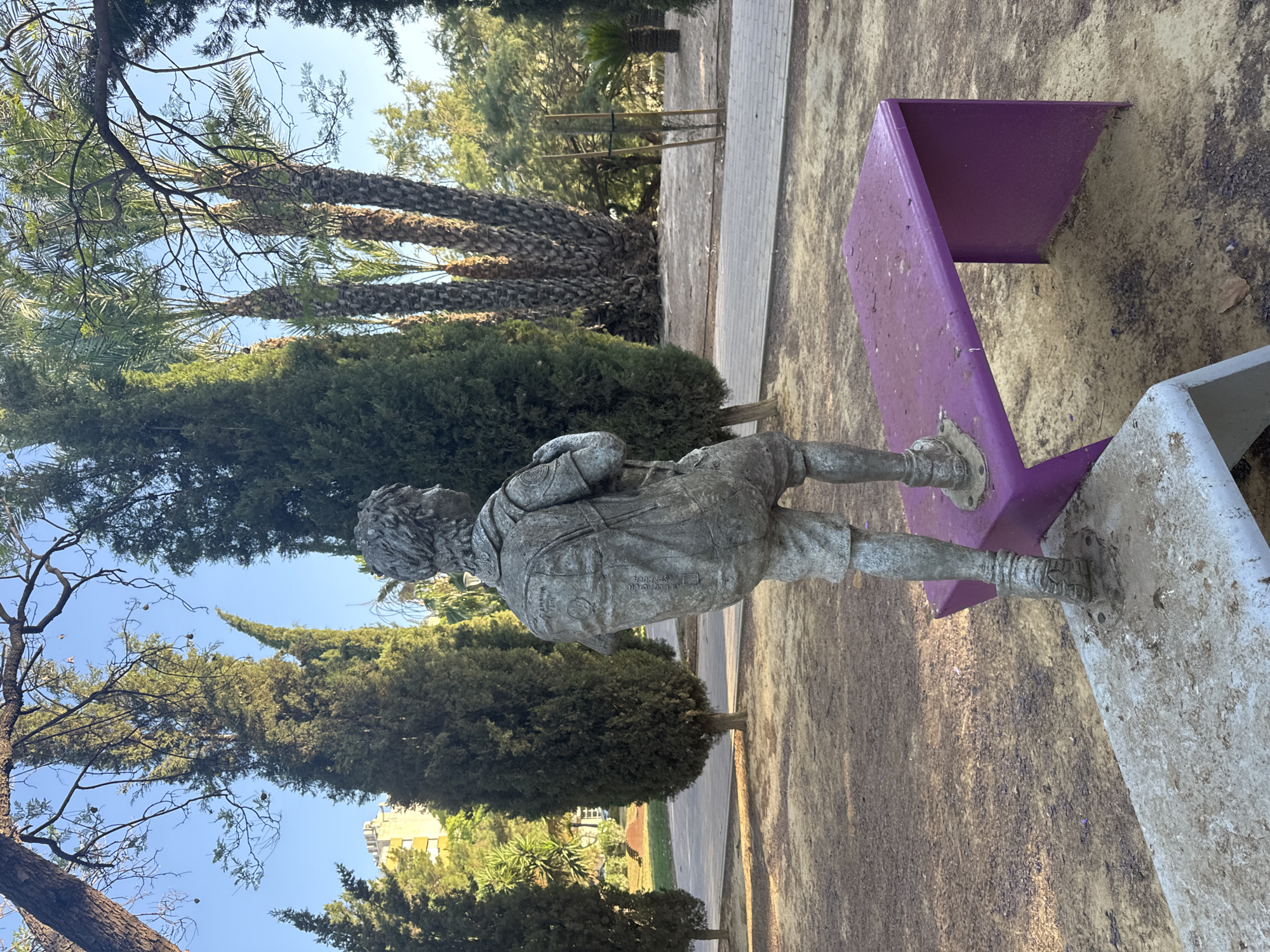 This image depicts a statue of a young boy wearing a backpack and shorts, standing on a purple bench in a park. The statue is made of a grey, weathered material and appears to be in motion, with one leg forward. The background features several tall cypress trees, palm trees, and some foliage, suggesting a Mediterranean climate. A paved pathway runs behind the statue and bench. In the distance, a yellow building is visible. The ground is covered in dirt and scattered leaves. This scene is located in the garden of the *Museo Salzillo* in the city of Murcia, Spain, a place known for its impressive collection of religious sculptures by Francisco Salzillo.