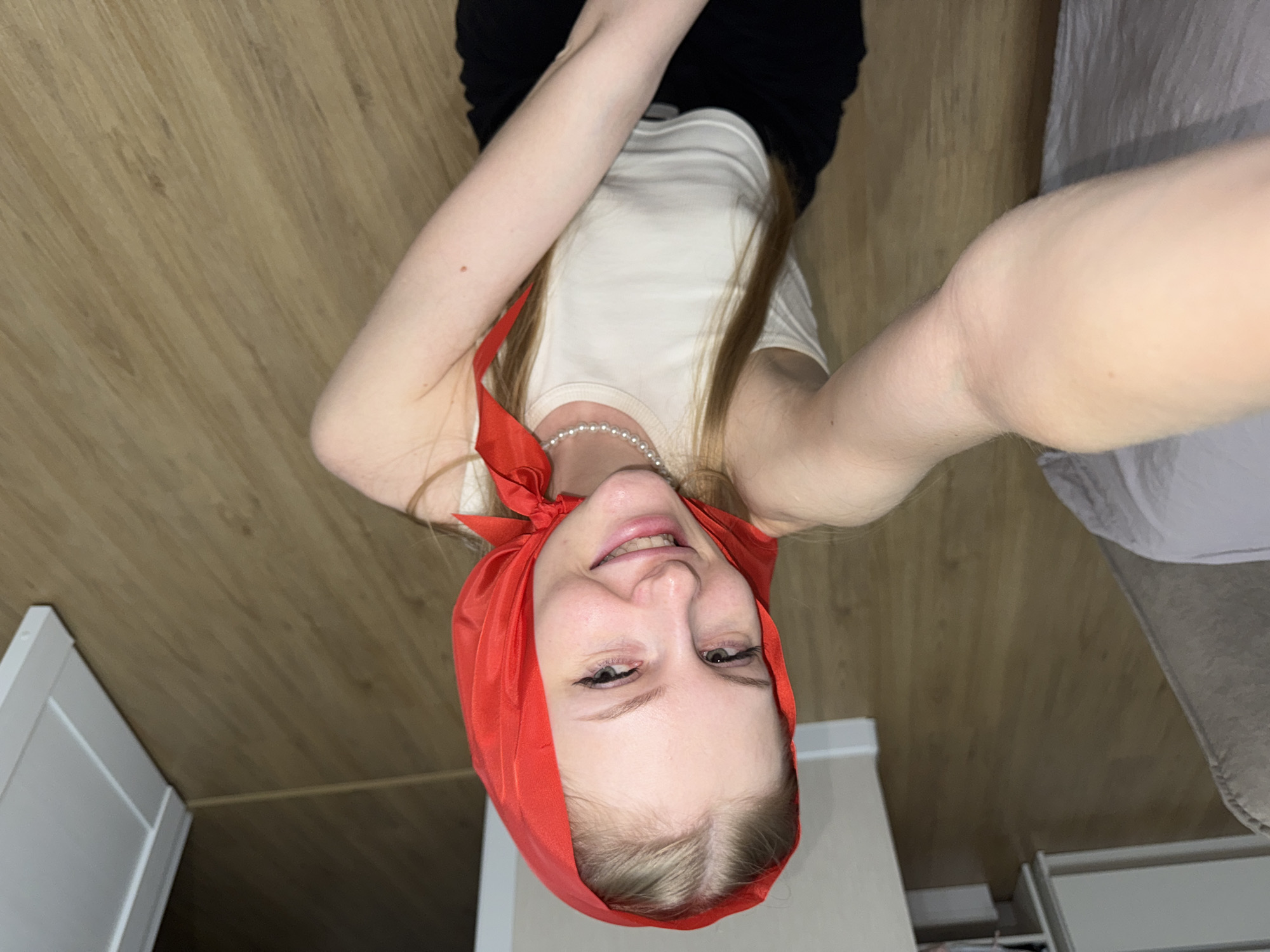 The image depicts a young woman lying on a wooden floor. She is wearing a white tank top, black pants, and a red headscarf tied at the back of her head. A pearl necklace adorns her neck. Her right arm is extended towards the camera, with her hand blurred. Her left arm is bent, resting on her chest. She is smiling and looking directly at the camera. The environment appears to be an indoor setting, possibly a room in an apartment, with a white piece of furniture visible in the background on the left. The wooden floor has a distinct grain pattern. There are no identifiable landmarks or elements within the image that would suggest a specific location in the city of Perm, Russia.