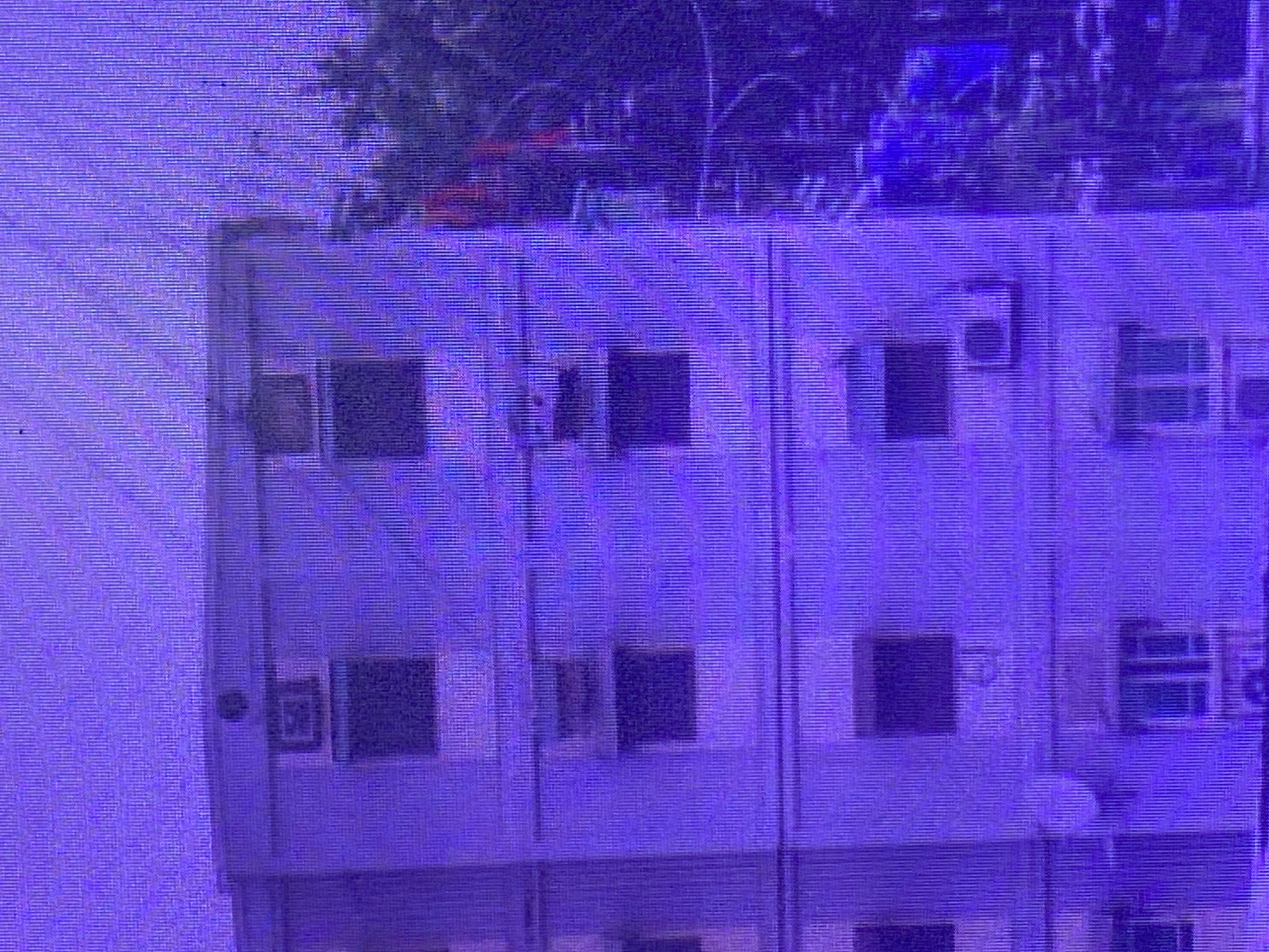 The image shows a close-up view of a multi-story building, likely an apartment complex or residential building, with a predominantly purple hue due to image processing or lighting. The building appears to have multiple windows on each floor, some of which are dark, suggesting they are closed or the interiors are not illuminated. There are visible air conditioning units protruding from some of the windows, indicating a warm climate.
At the top of the building, there are glimpses of greenery, possibly trees or other vegetation, adding a natural element to the urban scene. The overall impression is of a typical urban residential area in Ikorodu, Nigeria, under what could be daylight conditions, though the specific time of day is not definitively clear due to the color cast. There are no people or explicit activities visible in the frame. The building's facade is somewhat simple, with a regular pattern of windows and vertical divisions between sections of the building. There is no discernible text visible in the image. The image quality is somewhat grainy, with horizontal lines characteristic of older display technologies or compression artifacts.