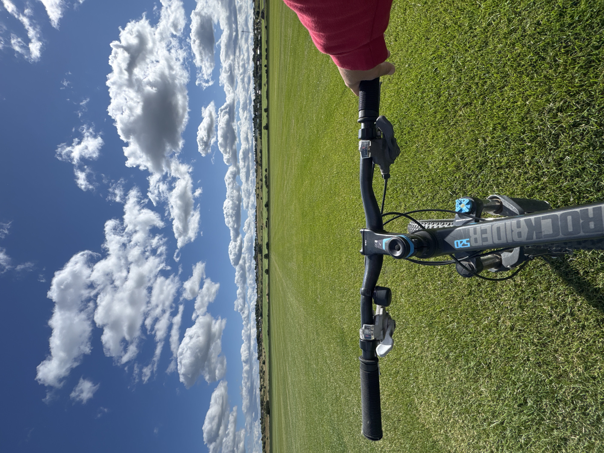 The image is a first-person perspective of someone on a bicycle, holding the handlebars, with a large, green, mowed field stretching out ahead. The field is bordered by a distant treeline and a small settlement of buildings. The sky above is bright blue with scattered white, fluffy clouds. The bicycle handlebars are visible in the foreground, with a red sleeve of a person's arm on the right side. The bicycle frame has "ROCKRIDER 520" printed vertically on it. The scene appears to be outdoors in a rural or semi-rural area. The specific location is not discernible from the visual elements alone.