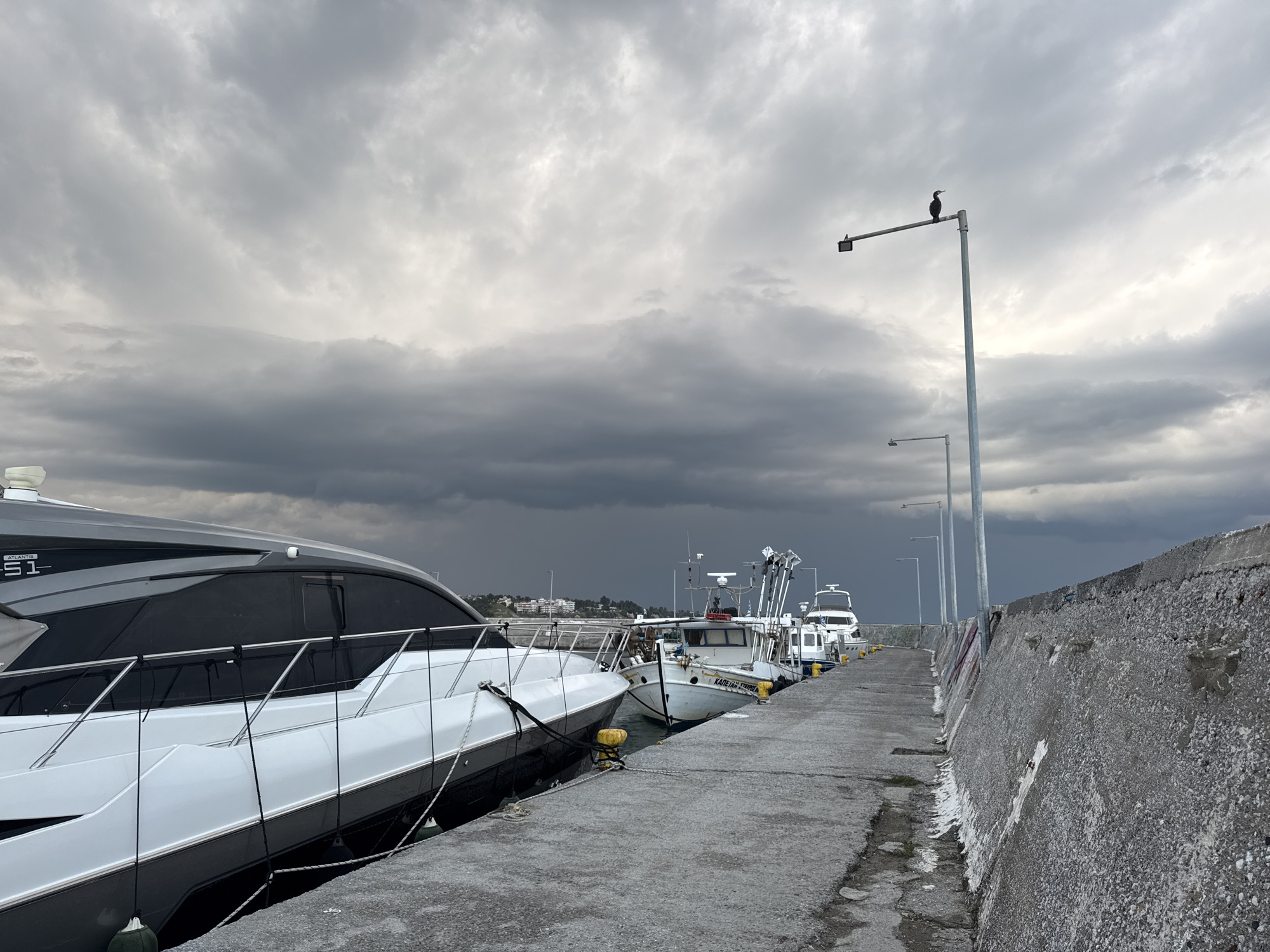 The image depicts a marina or harbor in Νέα Καλλικράτεια, Greece. Several boats are moored along a concrete pier. The largest boat on the left is a modern yacht with a gray and white hull. Further down the pier, smaller fishing boats are visible. A concrete seawall runs along the right side of the pier. Several lamp posts line the pier, with a bird perched on top of one. The sky is overcast with dark, heavy clouds, suggesting an approaching storm. The pier appears to be deserted.
