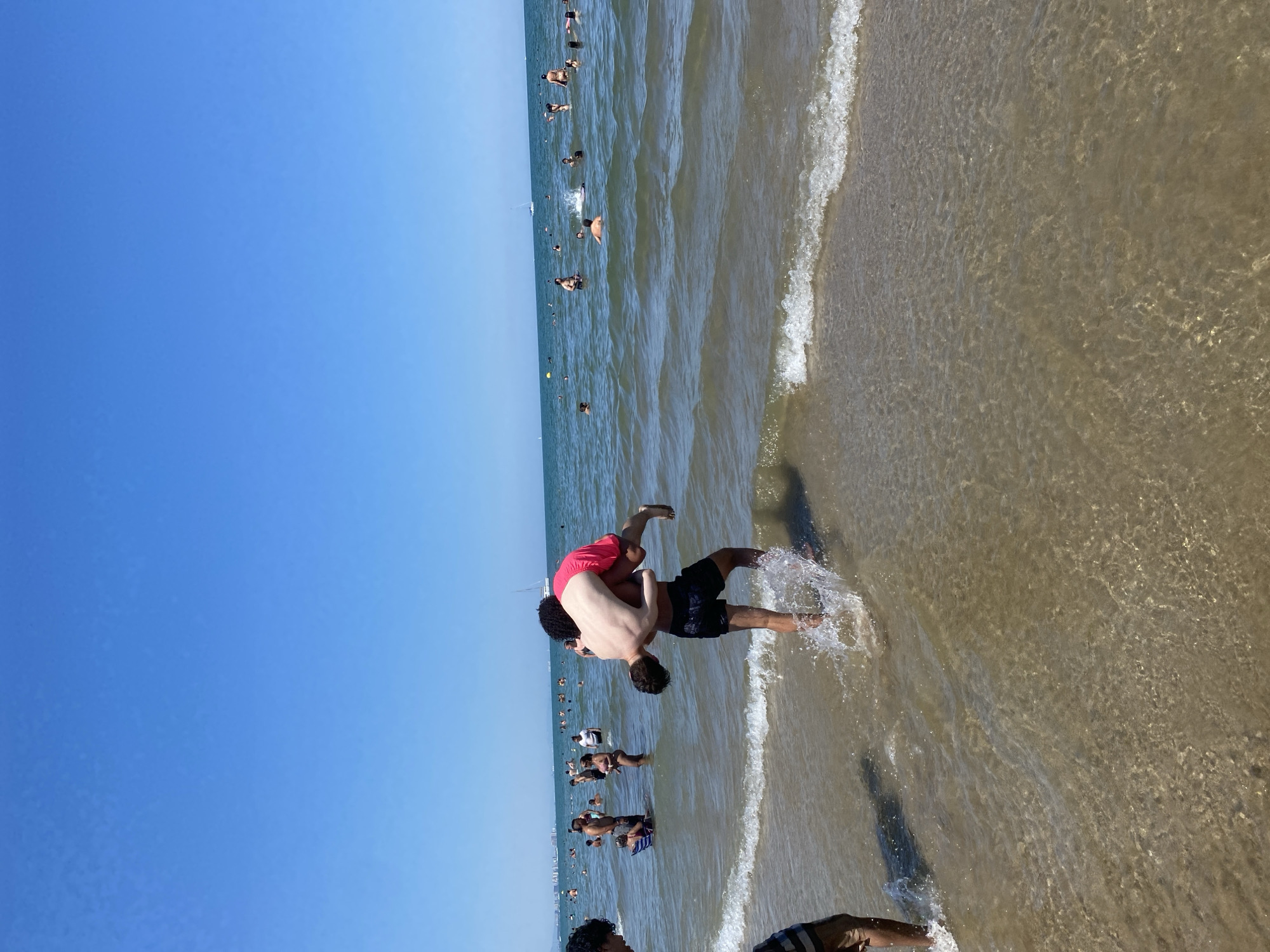 This image depicts a crowded beach scene on a sunny day. In the foreground, two young men are wading in the shallow water. One man, wearing black shorts, is carrying another man on his shoulders. The man being carried is wearing bright pink shorts. Both are generating splashes as they move.
In the mid-ground and background, the sea is populated with numerous other beachgoers swimming, standing in the water, or relaxing. The water is relatively calm with small waves breaking near the shore. The sky is clear and blue.
The overall scene is consistent with a typical summer day at a popular urban beach in Valencia, Spain, likely somewhere along the city's coastline such as Malvarrosa Beach or El Cabanyal Beach, known for their popularity and amenities. The presence of many people, the clear sky, and the type of beach environment strongly suggest this location.