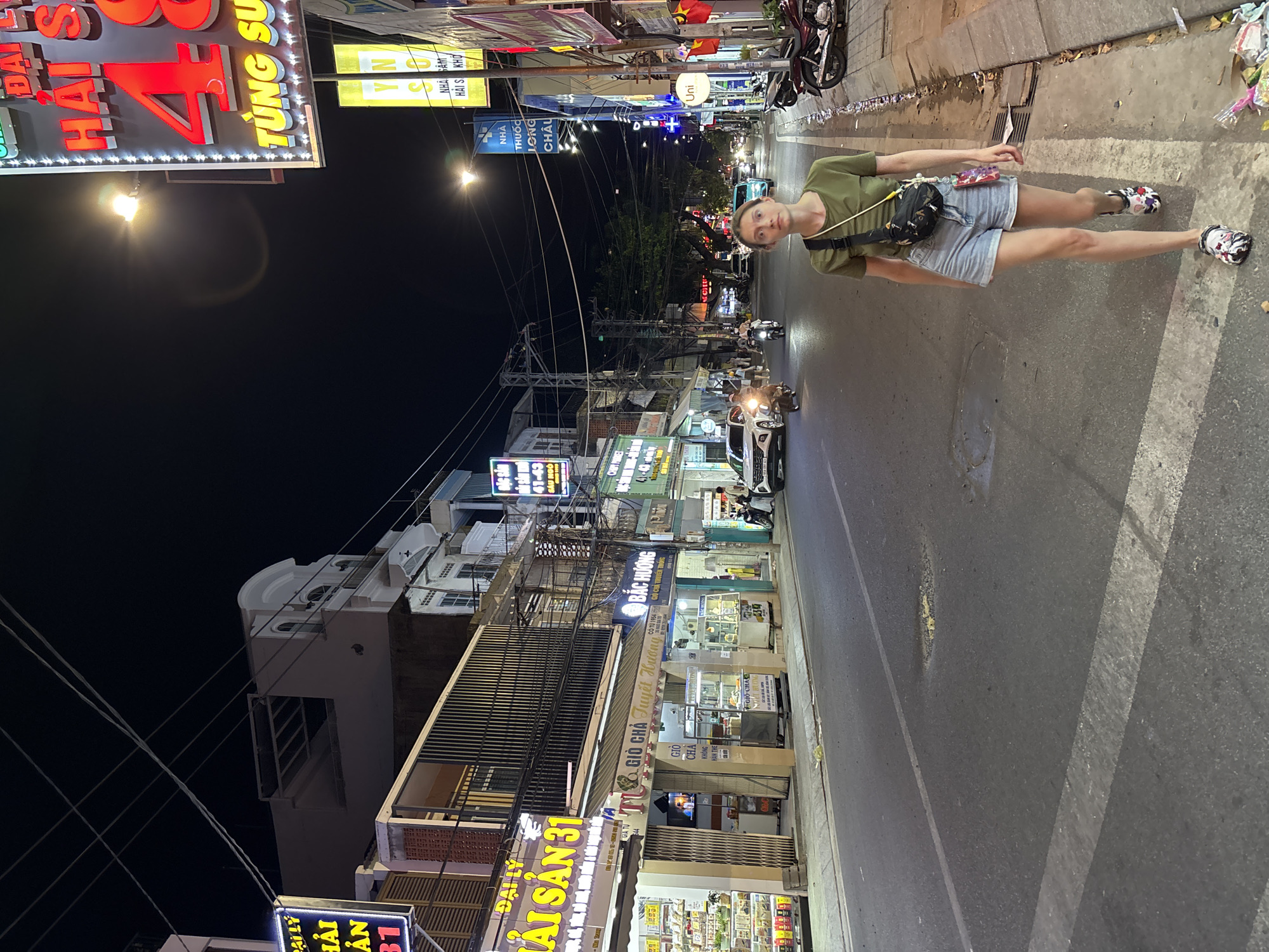 A medium shot shows a Caucasian woman walking on a dimly lit street at night in Nha Trang Ward, Vietnam. She is wearing a green t-shirt, denim shorts, and patterned clogs. A black crossbody bag with a colorful strap is slung across her body. The street is lined with shops and buildings displaying neon signs. In the background, vehicles and motorbikes are visible, and power lines crisscross overhead. The woman is looking towards the camera with a neutral expression. The scene appears to be a typical street scene in a Vietnamese city at night, with commercial establishments and traffic.