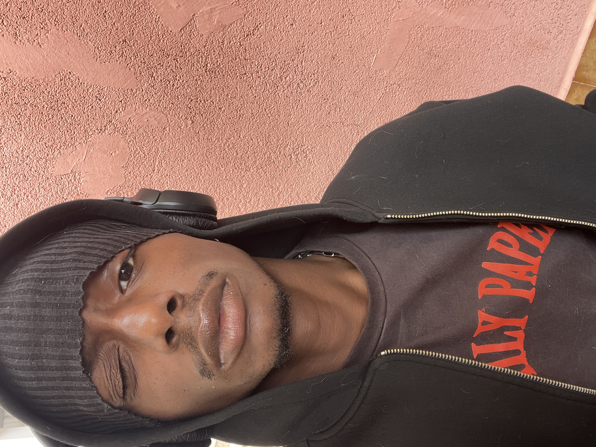 A vertical, head-and-shoulders portrait captures a young man of color against a textured, light reddish-brown wall. He is centered in the frame, looking directly at the viewer. He wears a dark, possibly striped, beanie or the hood of his jacket is pulled up over his head, partially covering his ears. A pair of black over-ear headphones rests around his neck, just below the beanie/hood.
The man is dressed in a black hooded zip-up jacket, unzipped to reveal a dark-colored t-shirt underneath. The t-shirt features red lettering, with "DAILY PAP" or "DAILY PAPER" clearly visible, though the full word "PAPER" might be partially obscured. His facial expression is casual; one eye is wide open and looking towards the camera, while the other is closed or squinted. He has a short, neatly trimmed mustache and a small patch of beard on his chin. His lips are slightly parted.
The background wall has a rough, stucco-like texture with subtle variations in color and relief, indicating a somewhat aged or rustic surface. Bright, even lighting illuminates the scene, suggesting it is daytime. A small hint of a lighter-colored surface, possibly a floor or different wall, can be seen at the bottom right corner of the image. The man's dark clothing shows some visible lint or light fibers. The image conveys a casual, perhaps candid, moment, set in what is presumed to be Barcelona, Spain.