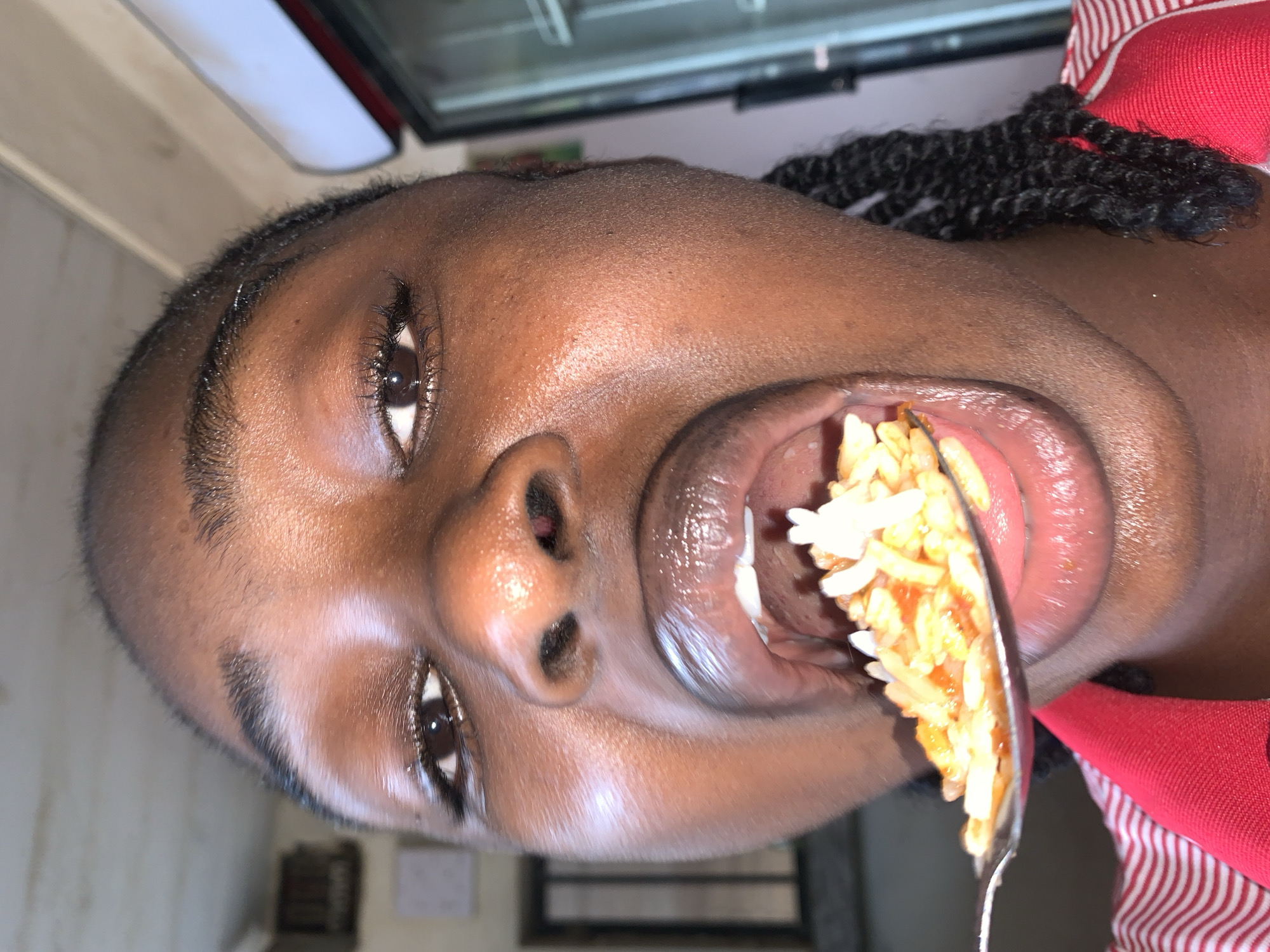 An close-up, rotated portrait-style image captures a person with dark skin in the act of eating. The image is oriented sideways, with the person's face filling the frame. A spoon laden with rice is positioned entering their widely open mouth.
The person has dark brown eyes, visible eyelashes, and short, dark hair or possibly braids partially visible towards the top right. Their skin appears smooth with some natural highlights. Inside their mouth, several white teeth are visible, along with the tongue. The rice on the spoon is a mix of white and reddish-orange grains, suggesting a dish like jollof rice. The spoon itself appears to be made of metal.
A garment in red with white stripes is visible on the right side of the frame, possibly part of a shirt or uniform. The background is out of focus and indistinct, suggesting an indoor setting with artificial lighting, though specific details are unclear. There is no visible text in the image. The moment captured is directly focused on the action of consuming food. Although the image provides no visual confirmation, the context specifies the location as New Stadium, Nigeria.