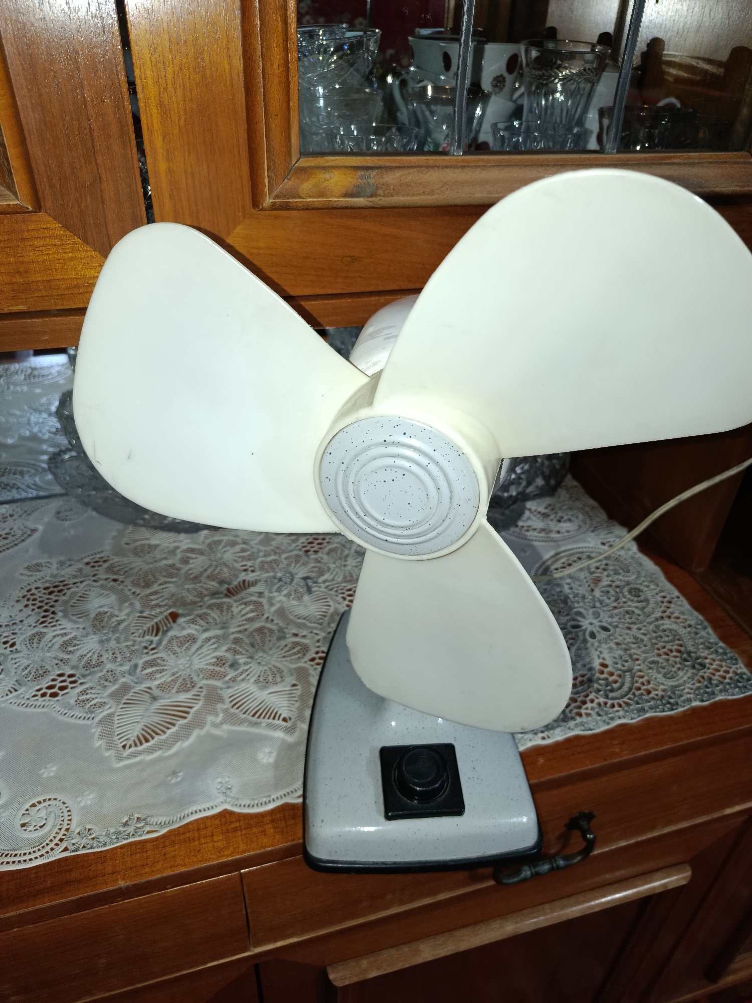 An old white electric fan with three blades sits on a wooden table covered with a white lace doily. The fan has a speckled gray central housing and a gray base with a black power button. The background shows a wooden cabinet with glass doors revealing a collection of glassware. The scene appears to be indoors, possibly in a home in Tashkent, Uzbekistan, given the context. There are no people present, and the fan is not in operation.