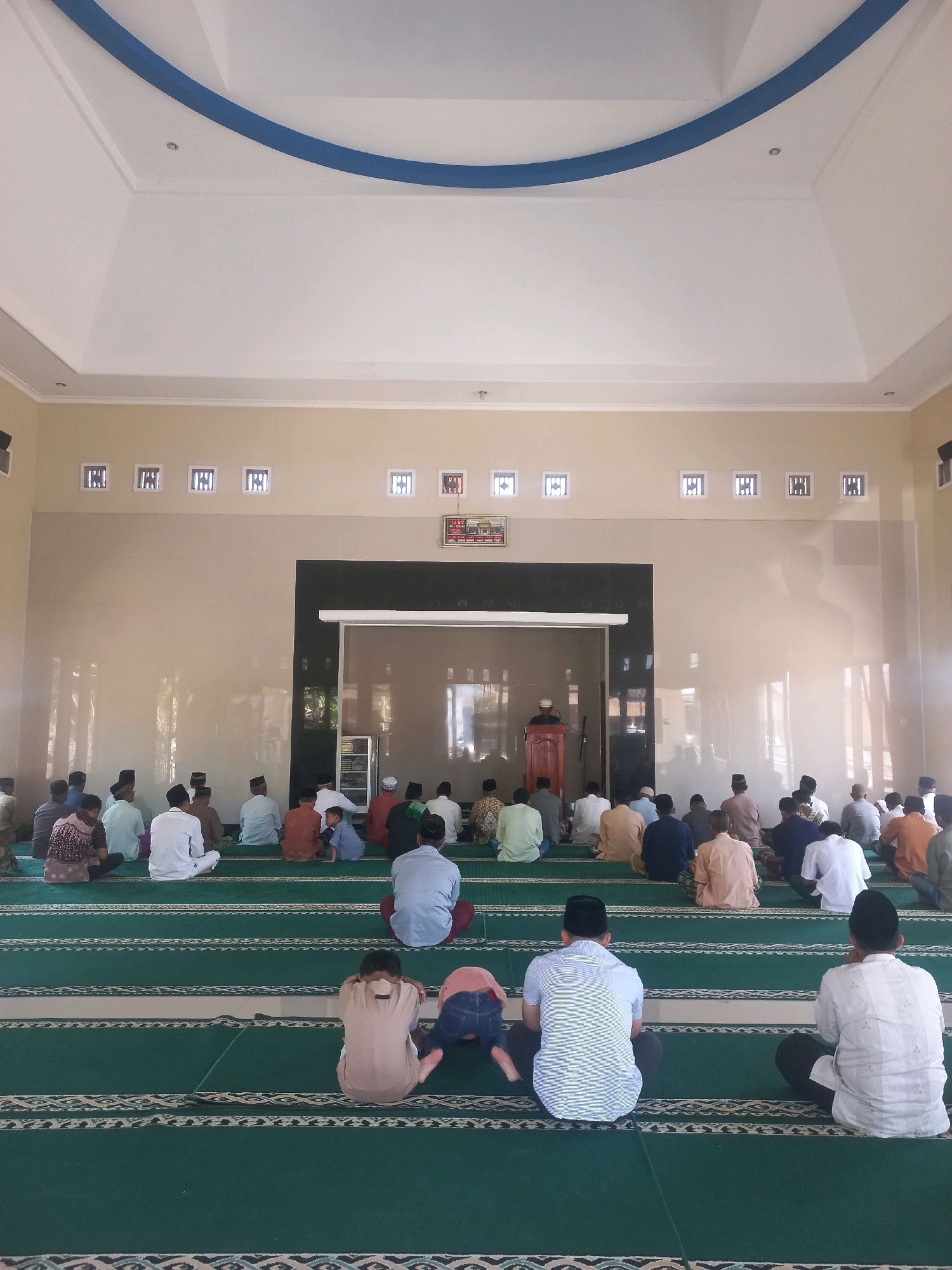 The image depicts the interior of a mosque in Nguntoronadi, Indonesia, during a prayer gathering. Approximately thirty men and boys are seated on a green carpeted floor, arranged in rows facing a dark partition wall. Some are in a seated position with their legs tucked beneath them, while others are in a kneeling or prostrating posture, indicating the ongoing prayer. In the center of the back wall, a man stands at a wooden podium, facing the congregation. The mosque features light-colored walls, a white ceiling with a blue decorative element, and several square windows with decorative grilles. A digital clock is visible on the right side of the back wall. The overall atmosphere appears to be one of communal worship and spiritual gathering.