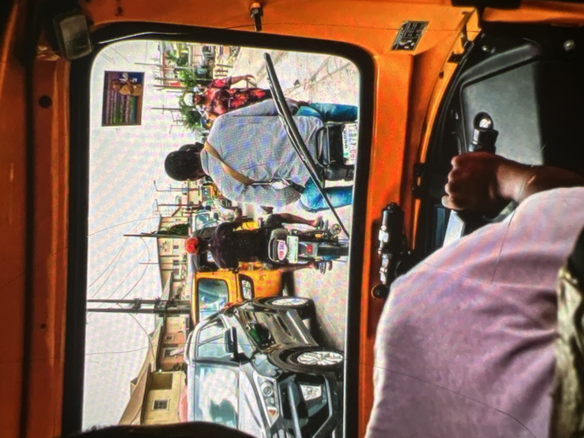 The image is a view from inside an open-air, orange-colored vehicle, likely an auto-rickshaw, looking out onto a busy street in Lagos, Nigeria. The foreground shows the arm and shoulder of the vehicle's driver, who is wearing a light pink garment and holding onto a steering mechanism.
The middle ground reveals the street scene. A black SUV is parked on the left, facing forward. Behind it, a yellow auto-rickshaw is visible. To the right of the SUV, a person wearing a red cap and a dark shirt is riding a motorcycle, facing away from the camera. Further down the street, another person is riding a motorcycle, wearing a light blue and white patterned shirt, jeans, and a light-colored head covering. A woman in a vibrant, multi-colored dress and a head covering is walking on the right side of the street.
In the background, there are buildings, some appearing to be residential or commercial, with houses having visible windows and rooftops. Power lines and poles are also present, typical of urban infrastructure in Lagos. A small framed advertisement or sign is attached to a pole on the right side of the street. The overall impression is of a congested urban environment with active street life.