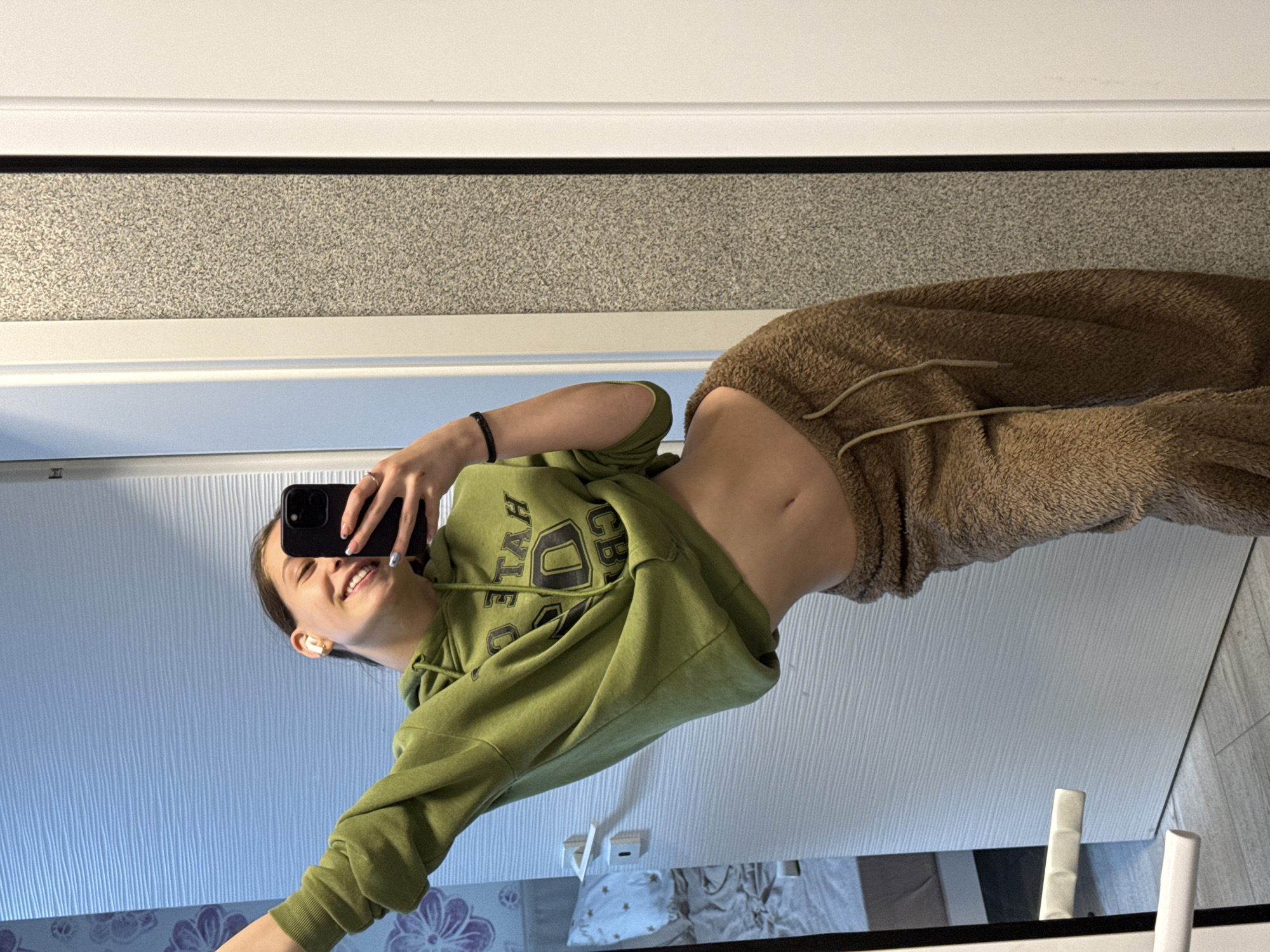 A young woman stands in a bathroom, facing a mirror and holding a black smartphone up to take a selfie. She is wearing a green hooded sweatshirt and brown sweatpants that are pulled down to expose her stomach. The bathroom has a speckled white and grey countertop above the mirror, a white door or wall below, and possible floral wallpaper near the bottom of the reflection. Additional objects visible in the mirror include what appears to be a bed, bedside tables, and bathroom fixtures. The city and specific location in Sofia, Bulgaria cannot be determined from the image.