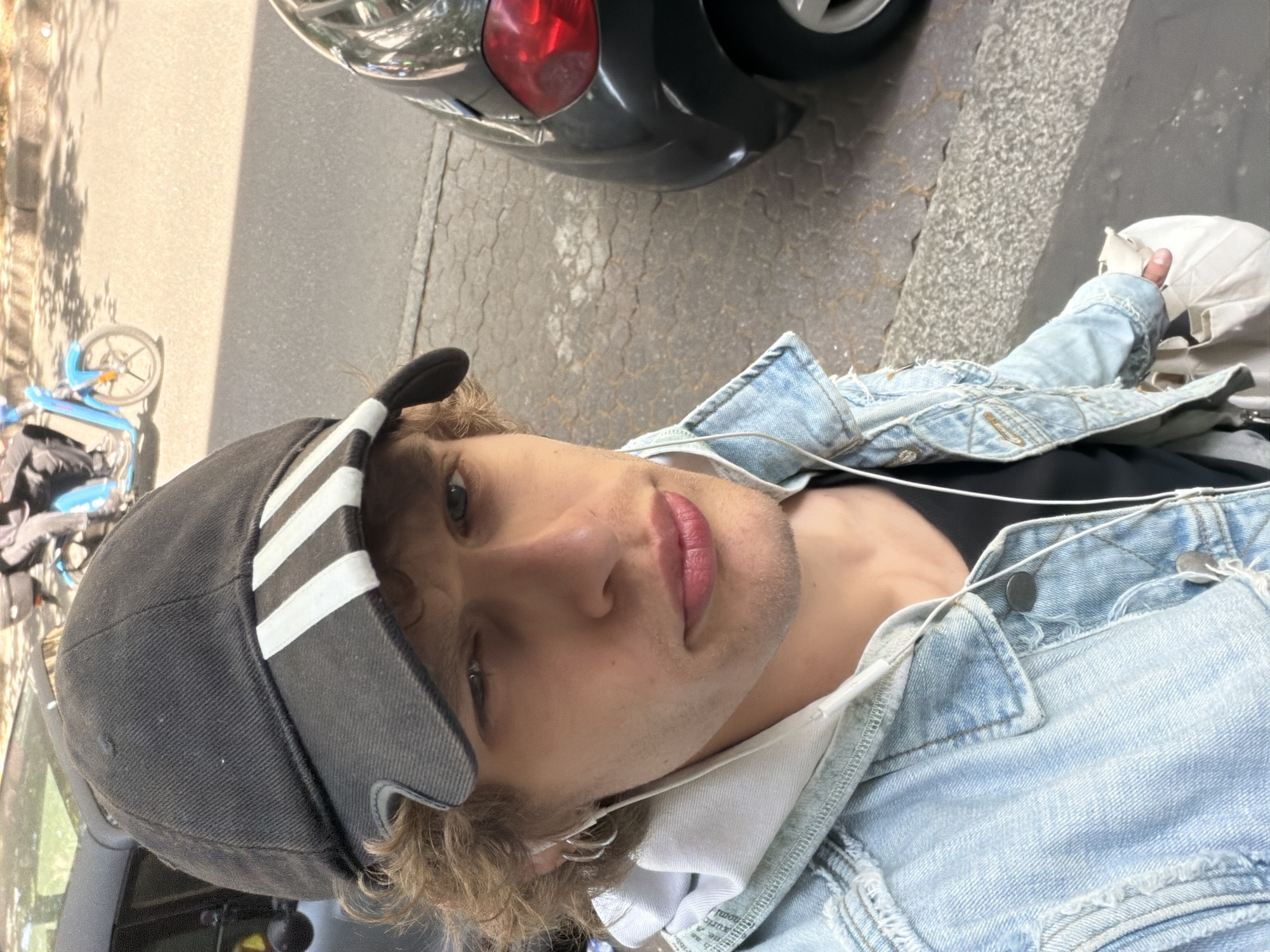 A young adult male stands on a paved surface in Milan, Italy, wearing a black baseball cap with white stripes, a light blue denim jacket over a dark top, and white headphones. He is positioned near a parked car, visible in the background, and a bicycle sharing station can be seen on the left side of the frame. The setting appears to be an urban street, likely during daylight.