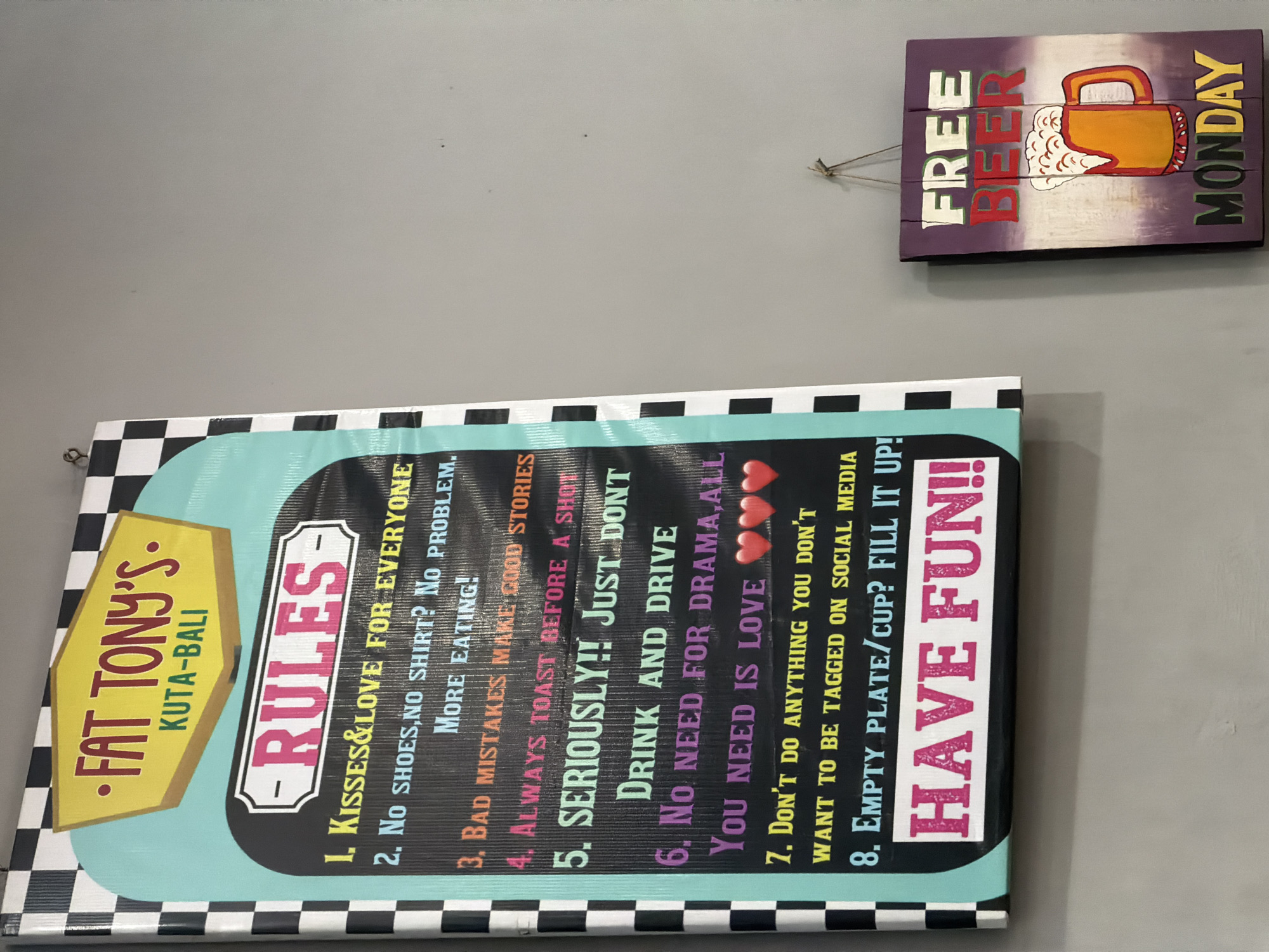 The image depicts two signs hanging on a gray wall, likely inside a bar or restaurant in Kuta, Bali, Indonesia.
The larger sign on the left is a vertical, rectangular board with a black and white checkered border. At the top, a yellow, hexagonal shape displays "FAT TONY'S" in red, and "KUTA-BALI" in yellow. Below this, a teal-colored curved shape frames the word "RULES" in large, pink, block letters. Beneath "RULES," a list of eight rules is written in various colored fonts:
1. Kisses & Love for Everyone
2. No shoes, no shirt? No problem. More eating!
3. Bad mistakes make good stories
4. Always toast before a shot
5. SERIOUSLY!! Just dont drink and drive
6. No need for drama, all you need is love (with three red heart emojis)
7. Don't do anything you don't want to be tagged on social media
8. Empty plate/cup? Fill it up!
The bottom of the large sign features a wide, horizontal pink band with the words "HAVE FUN!!" in large, white, distressed block letters.
To the right of the main sign is a smaller, vertical wooden sign with a purple background. It reads "FREE" in white letters at the top, followed by "BEER" in large red letters. Below this is a depiction of a frothy beer mug in orange and yellow hues. The bottom of this sign says "MONDAY" in yellow letters. The sign is hung by a thin string.
The overall scene suggests a casual and fun establishment. The lighting is even, and the wall appears to be a plain, unadorned surface. No people are visible in the image.