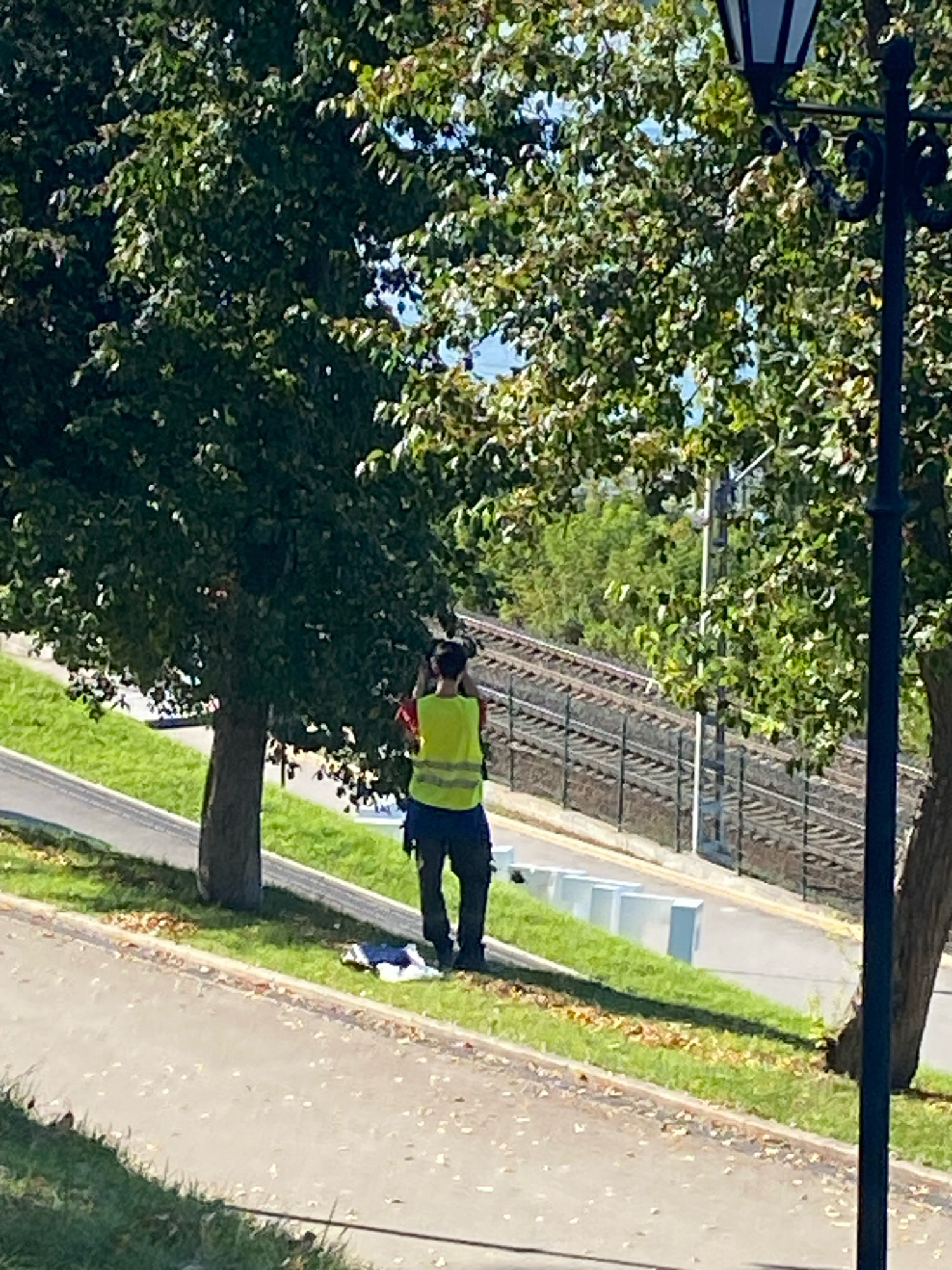 A person wearing a high-visibility vest is standing on a grassy slope near a paved walkway. In the background, a set of train tracks runs parallel to a fence, with trees and a lamppost also visible. The scene appears to be outdoors in a park or public area. The image does not contain enough information to determine the specific location in the city of Perm, Russia, or any further details about the individual or their actions.