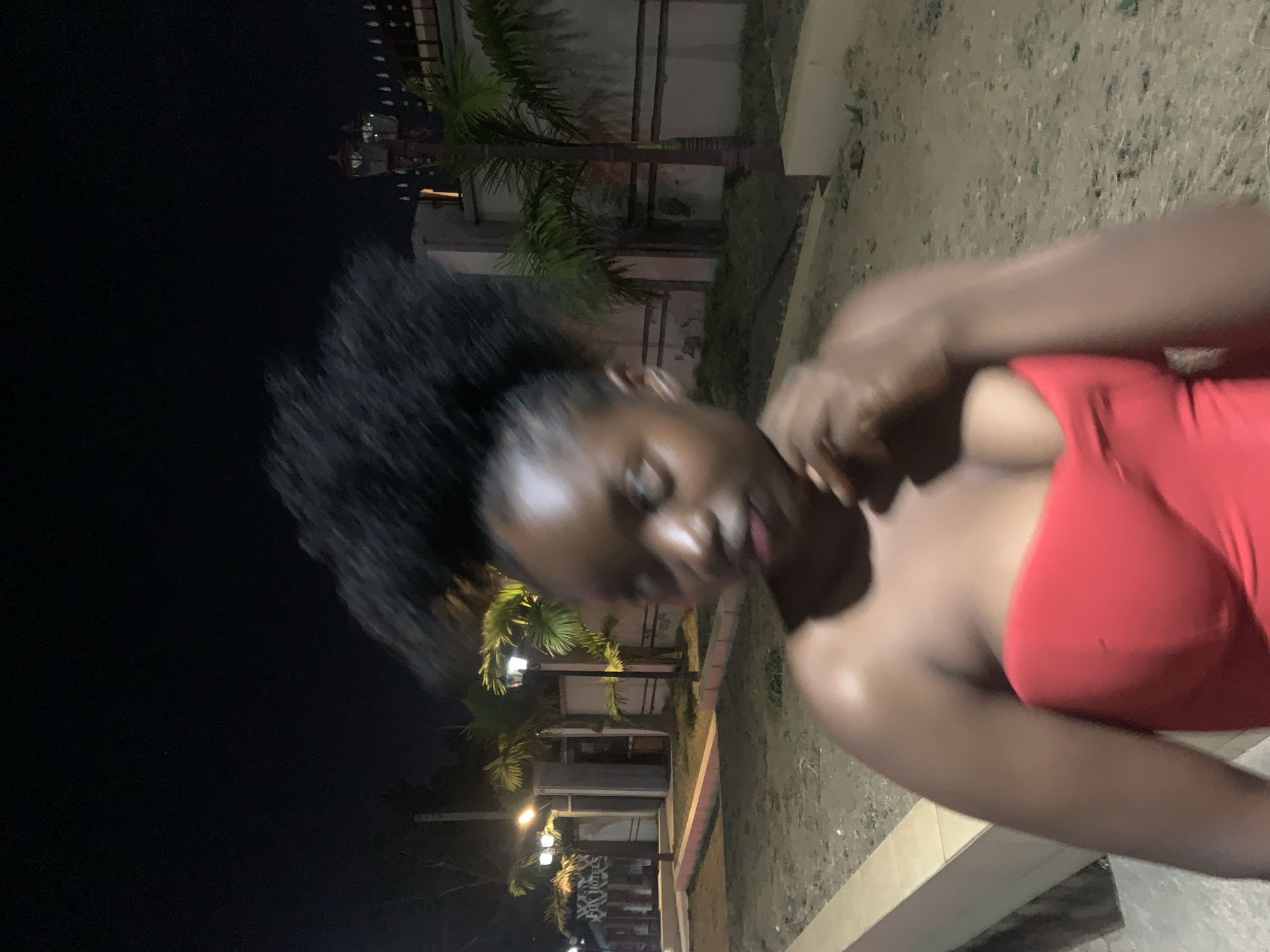 The image shows a woman with dark skin and black hair, wearing a red top, standing in an outdoor area at night. The location appears to be in Jos, Nigeria. In the background are palm trees and a building with lit windows. The ground appears to be concrete or pavement. The lighting is dim, primarily from streetlights.