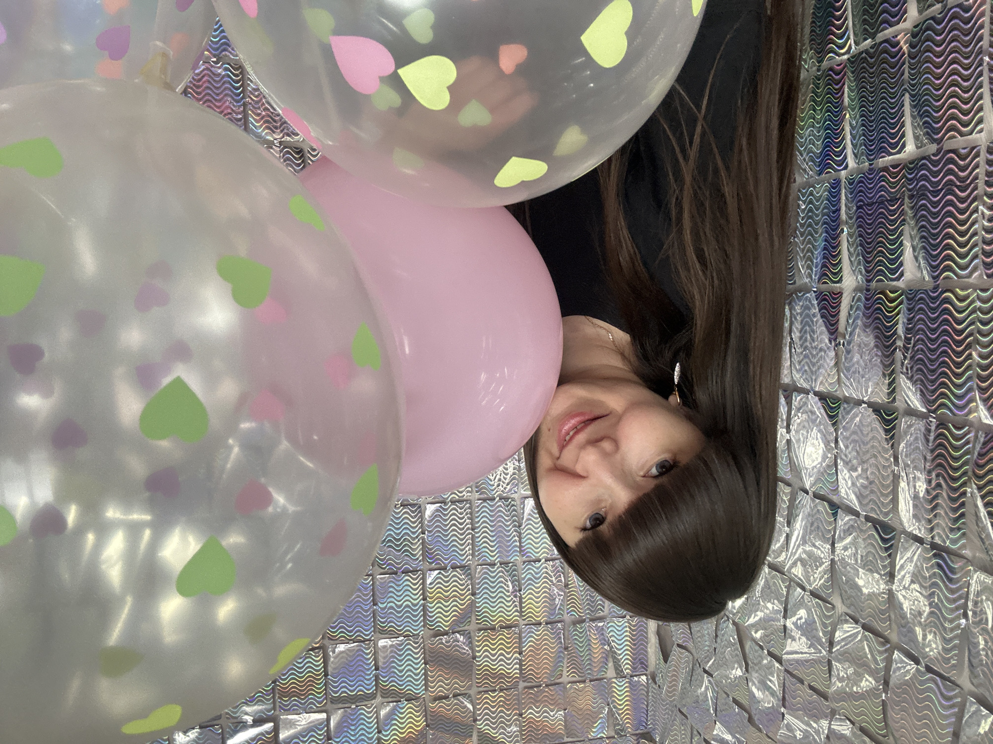 A woman with brown hair and bangs smiles, partially obscured by three balloons, a pink one and two clear balloons with colored heart patterns. She is wearing a black top and an earring is visible. She is in front of a reflective, shimmering wall composed of small square tiles, each with a wavy pattern. This image was taken in the city of Chaikovsky, Russia.