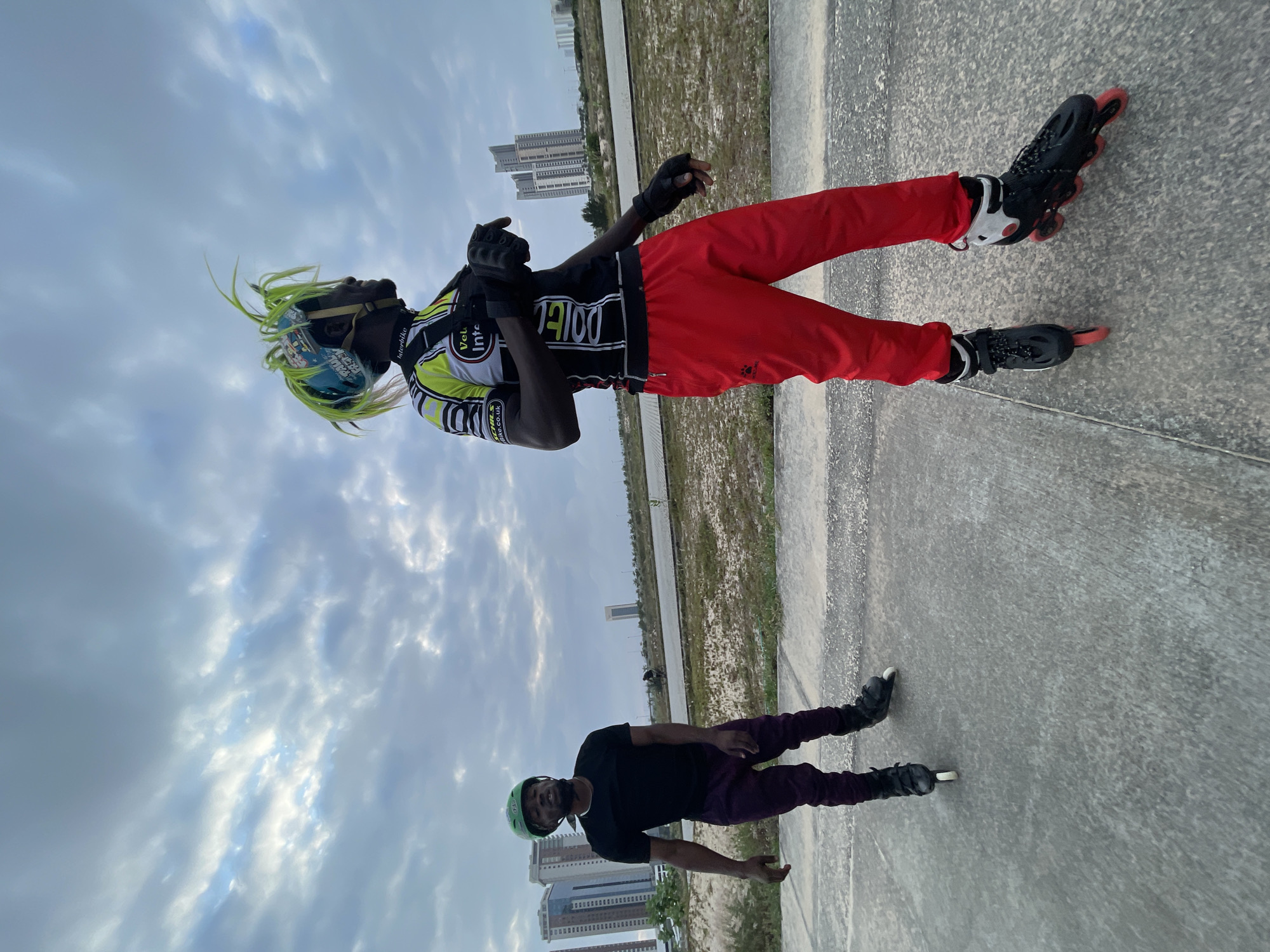 In Ilado, Nigeria, two individuals are engaged in inline skating on a paved outdoor track under a cloudy sky. The person in the foreground, facing right, wears a bright yellow and black cycling jersey with red pants and black inline skates. Their helmet is adorned with vibrant green faux hair. To the left, another individual, facing the camera, wears a black t-shirt, purple pants, and green inline skates, with a green helmet. In the background, modern high-rise buildings rise against the horizon, indicating an urban setting. The ground is a light-colored concrete path, bordered by dry vegetation and a dividing line. A distant, silhouetted figure on skates can be seen further down the path, suggesting a shared recreational space.