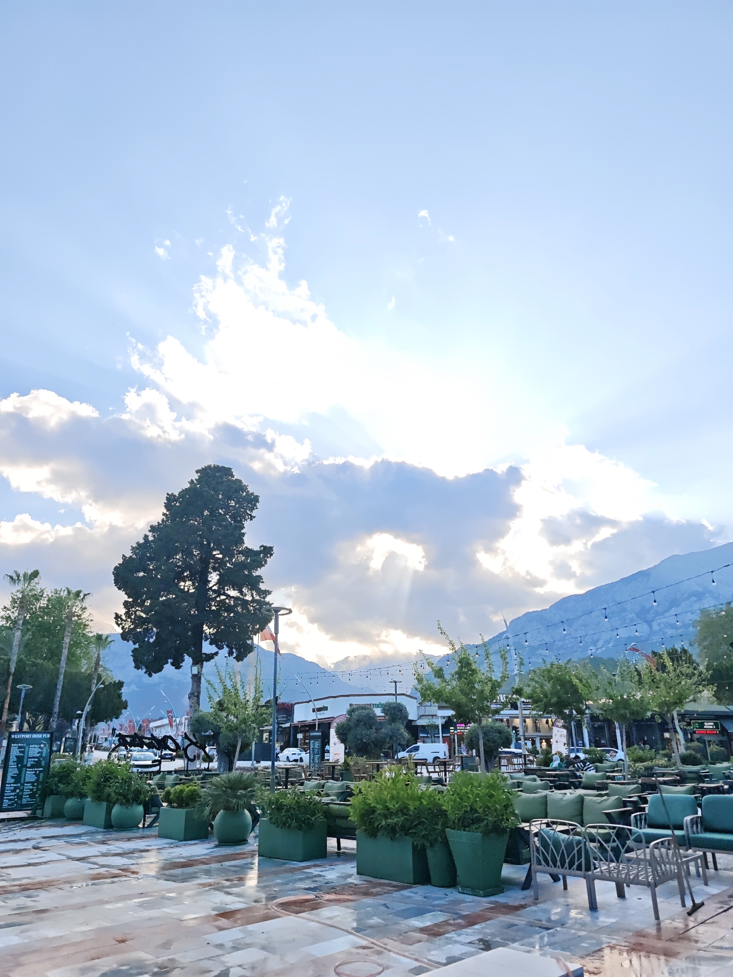 The image depicts an outdoor public space in Kemer, Turkey. A large tree stands on the left side of the frame. In the foreground, there are square green planters lining the outdoor seating area of a pub with green furniture. Beyond the planters, there's a paved area. Further back, there is a street with cars and buildings in the background. Mountains are visible in the far distance. String lights are hung overhead. The sky is bright, with the sun shining through the clouds.
